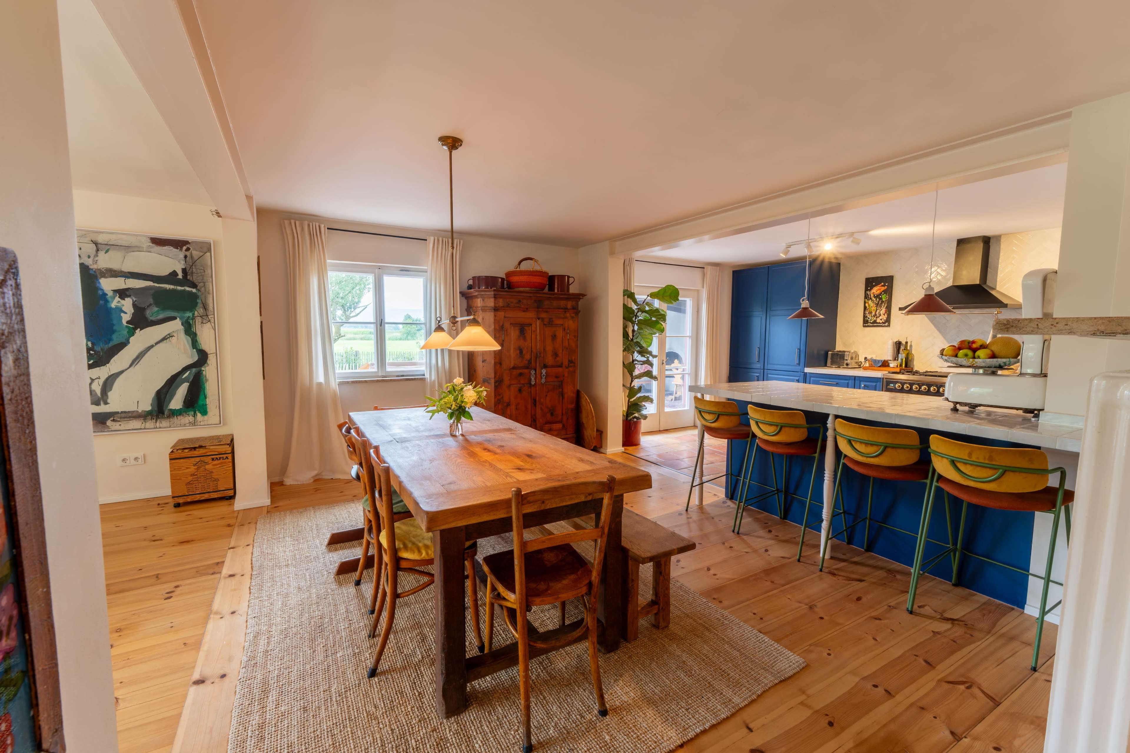 The image shows a spacious kitchen and dining area with a wooden dining table, colorful bar stools at an island, and large windows allowing natural light.