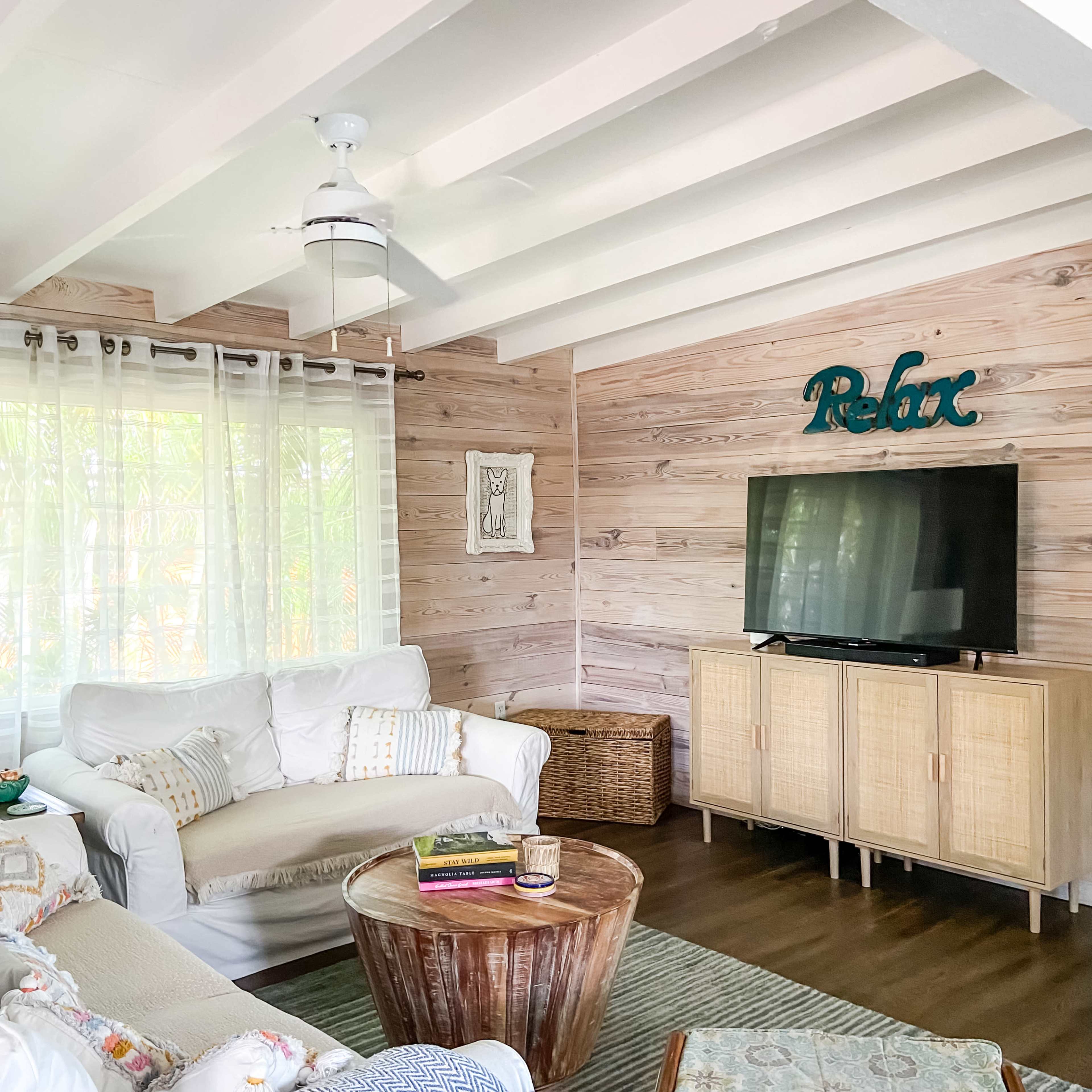 The room features a light-filled space with wooden walls, a white sofa, a wooden coffee table, and a television stand.