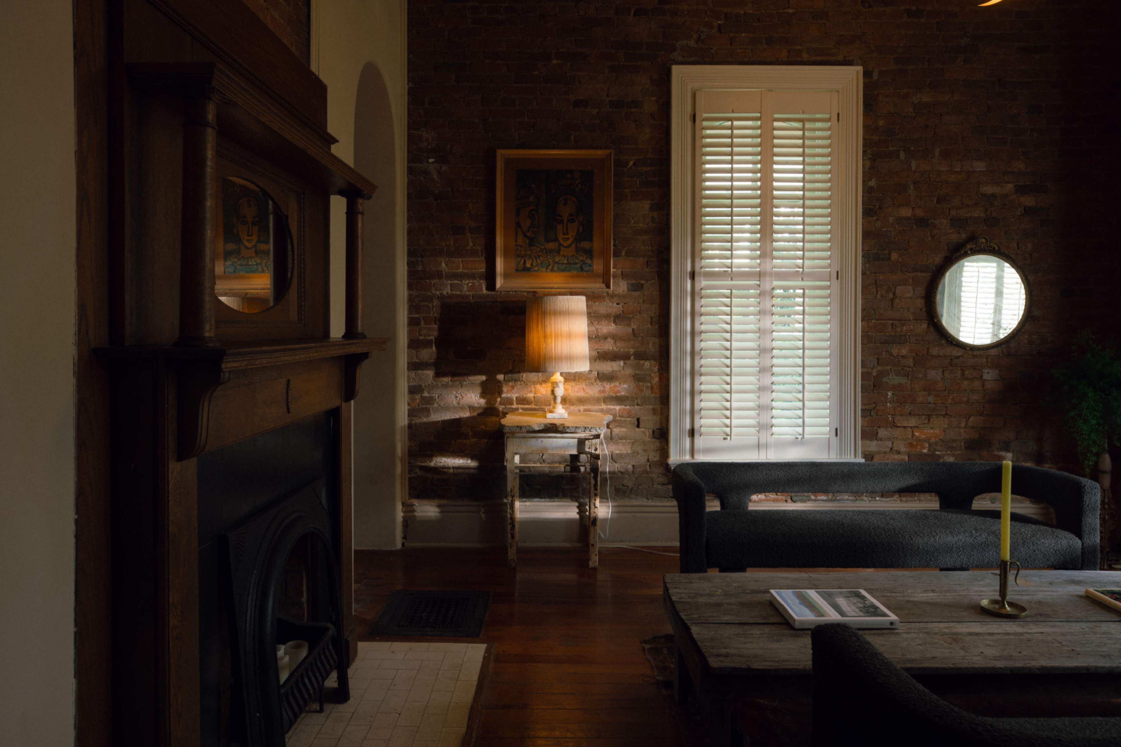 The room features a brick wall, a fireplace with a wooden mantel, a lamp on a side table, a large window with shutters, and a round mirror reflecting light.