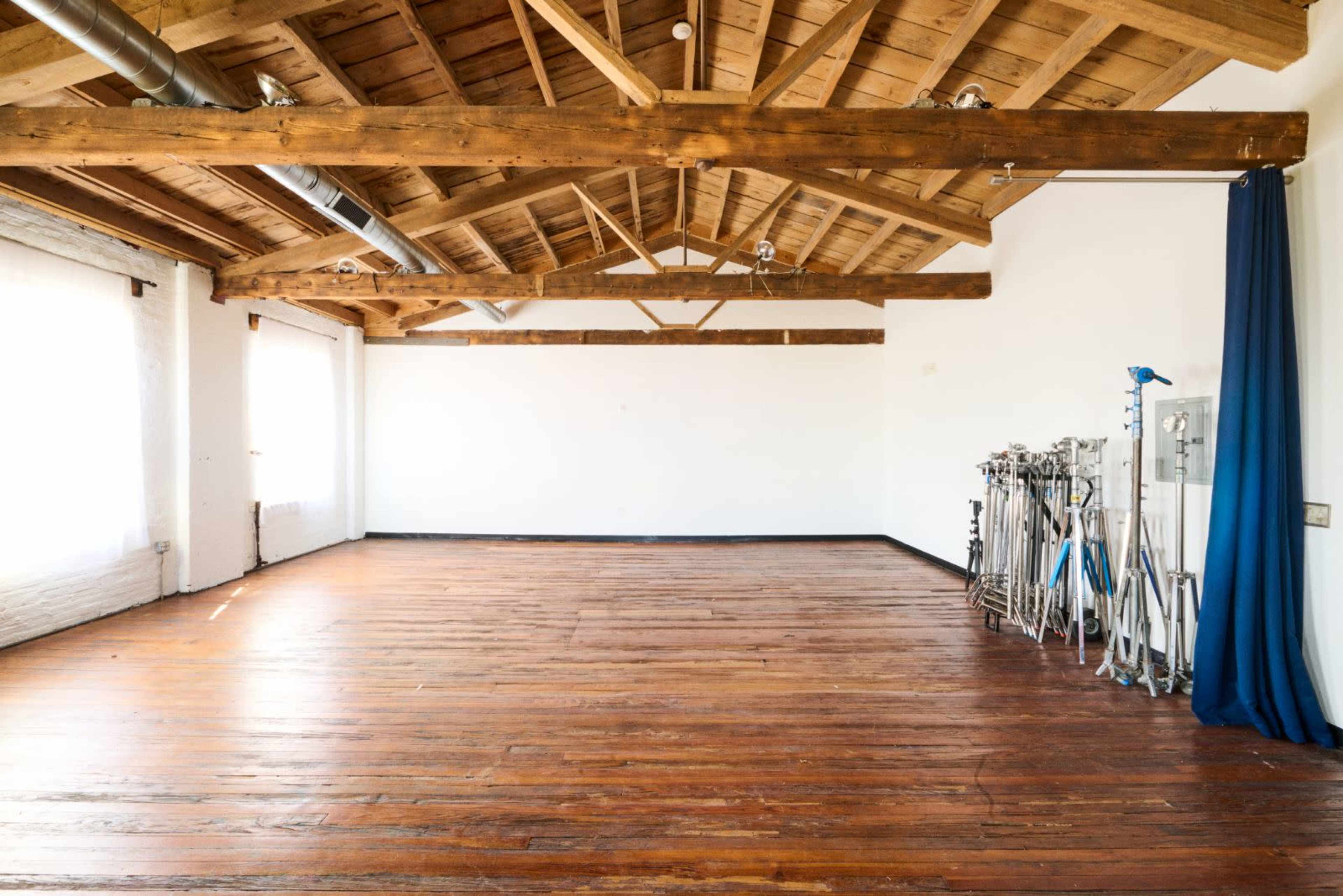 A spacious room with wooden beams and a polished wooden floor, featuring large windows and equipment stands along one wall.