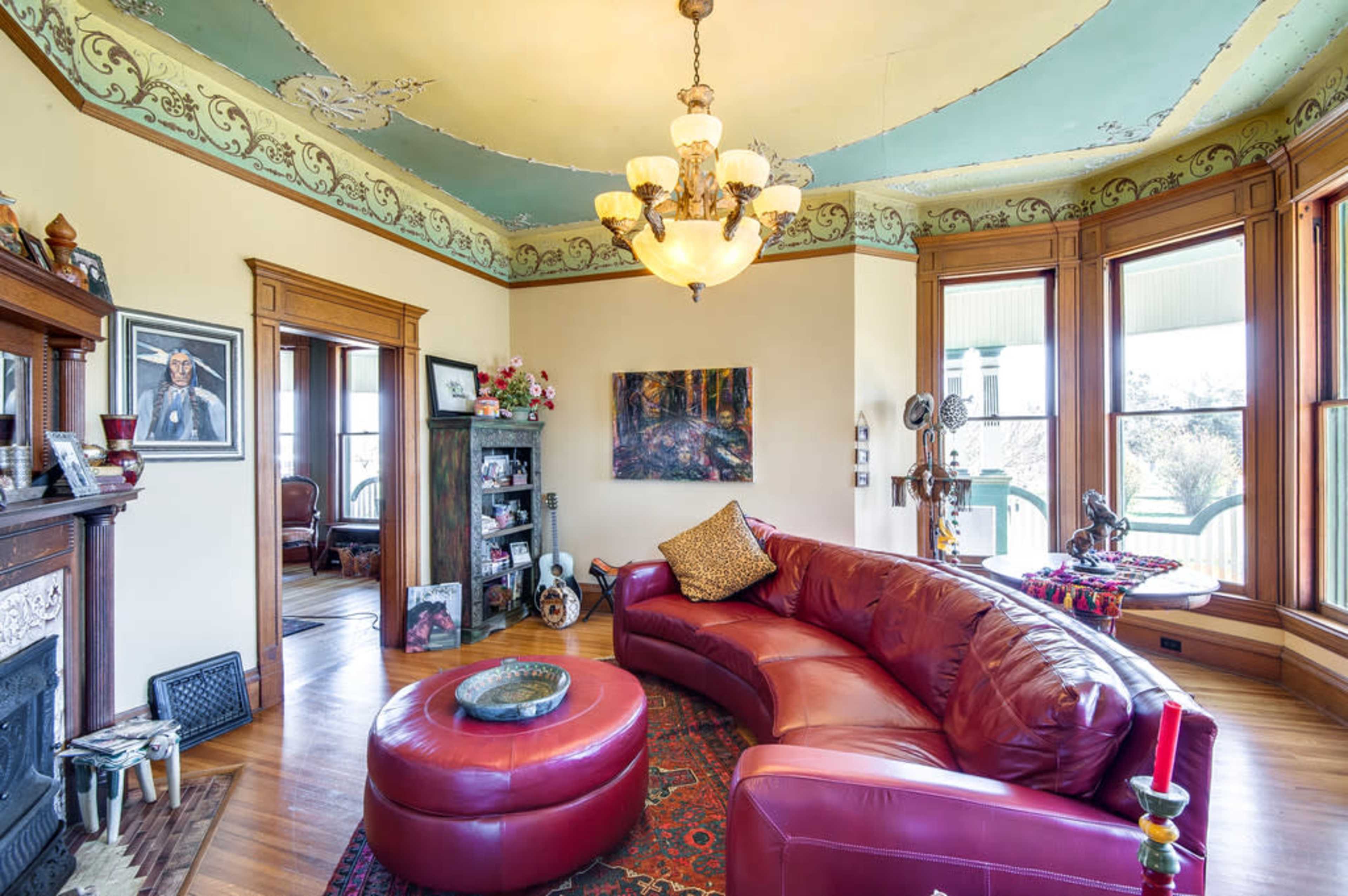 A cozy living room with a curved red leather sofa, a decorative ceiling, and a large chandelier, featuring wooden trim and large windows.