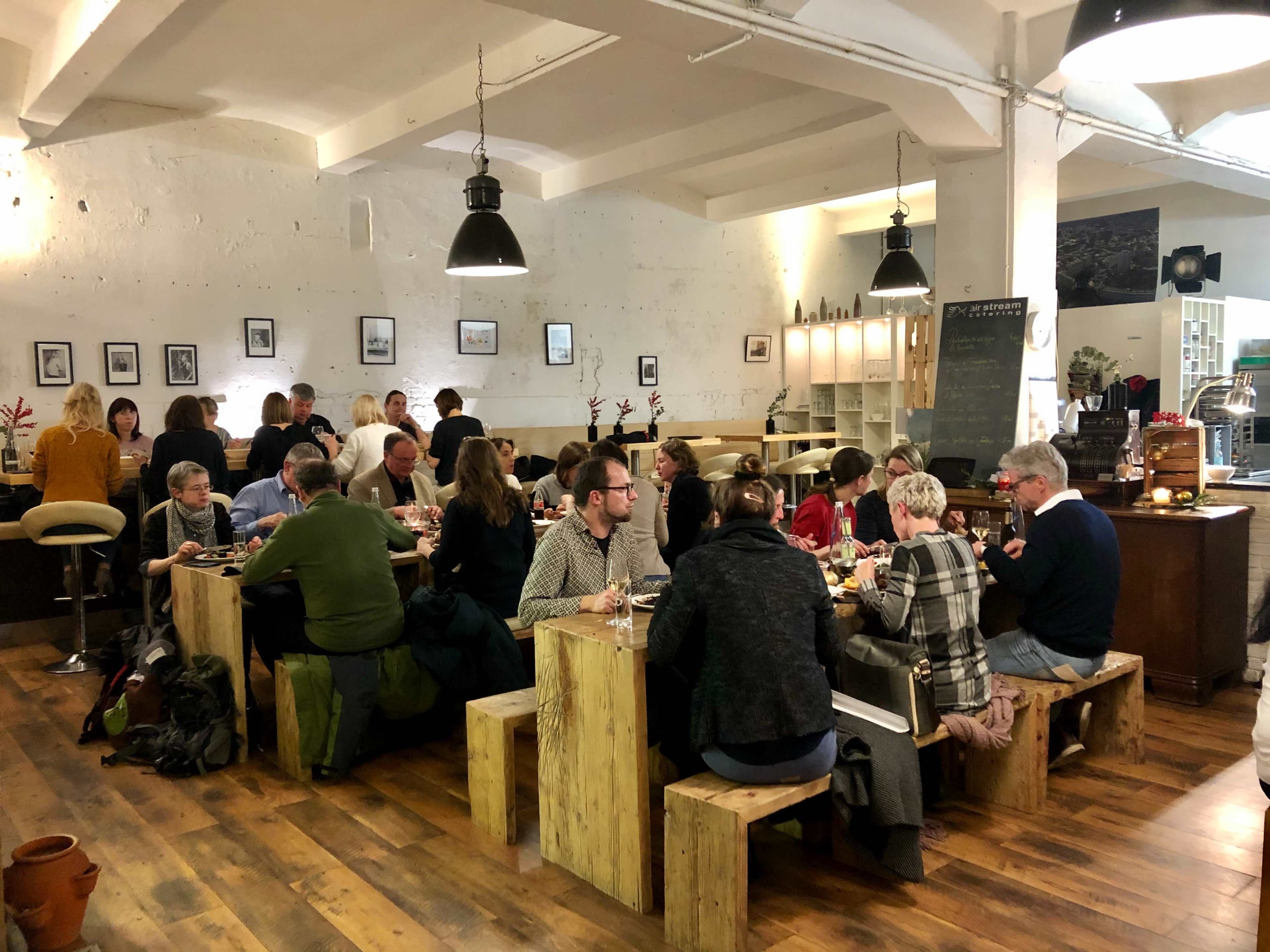 A group of people is seated at long wooden tables in a bustling restaurant with industrial decor, enjoying a meal together.