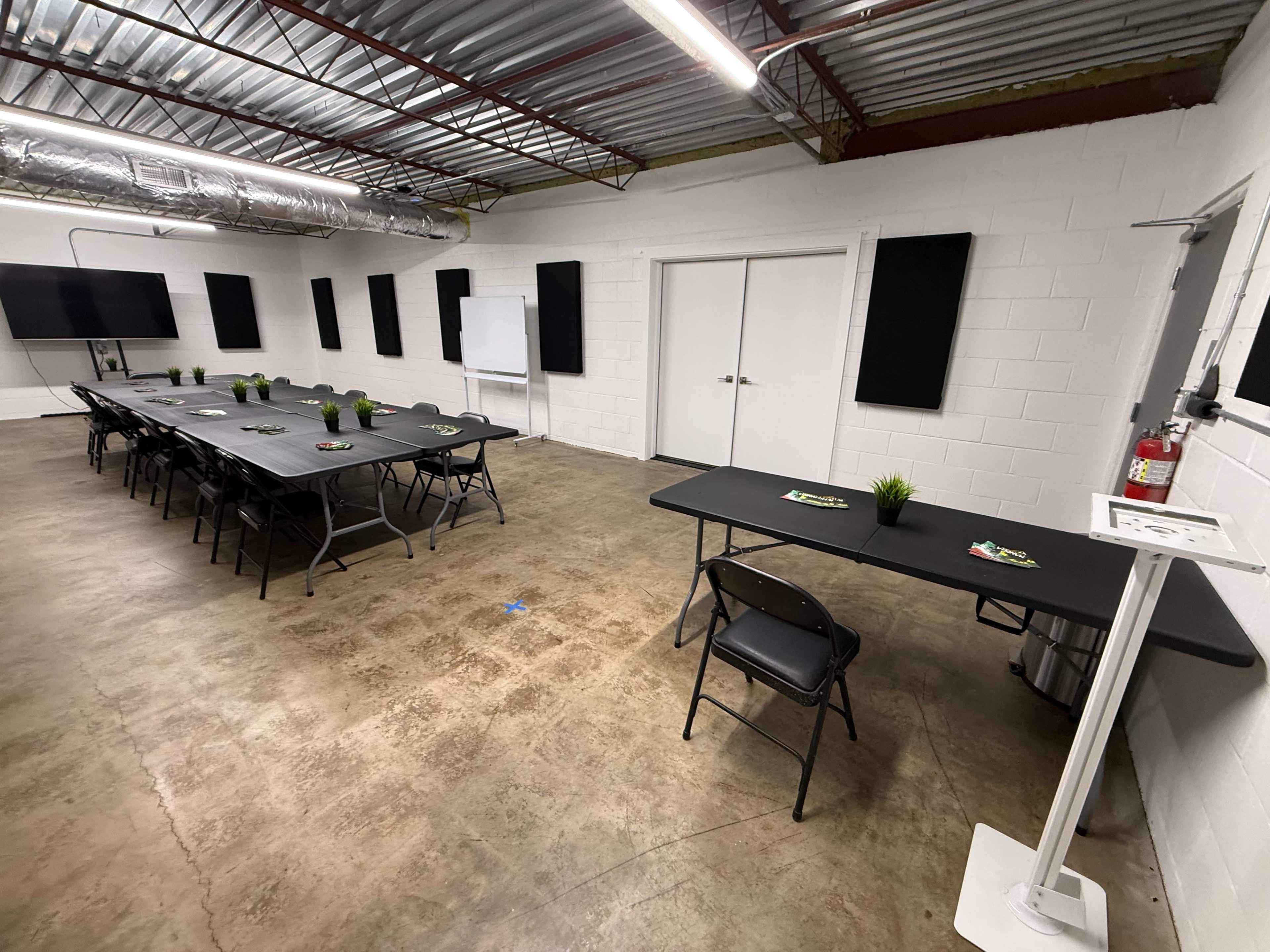 The image shows a large, empty meeting room with rows of tables and chairs, a whiteboard, and several plant decorations along the tables in a well-lit environment.