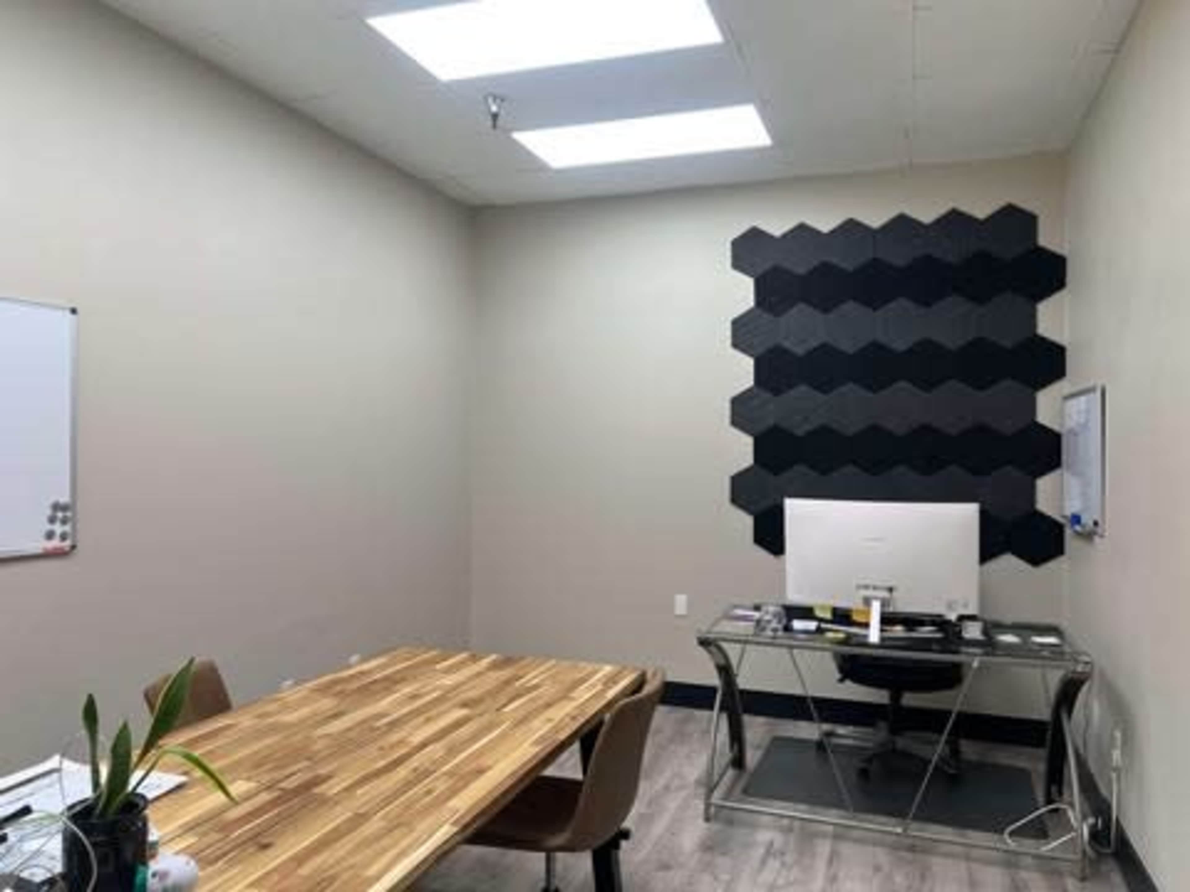 The image shows a minimalistic office space featuring a wooden table, a desk with a computer, and a textured black wall installation.