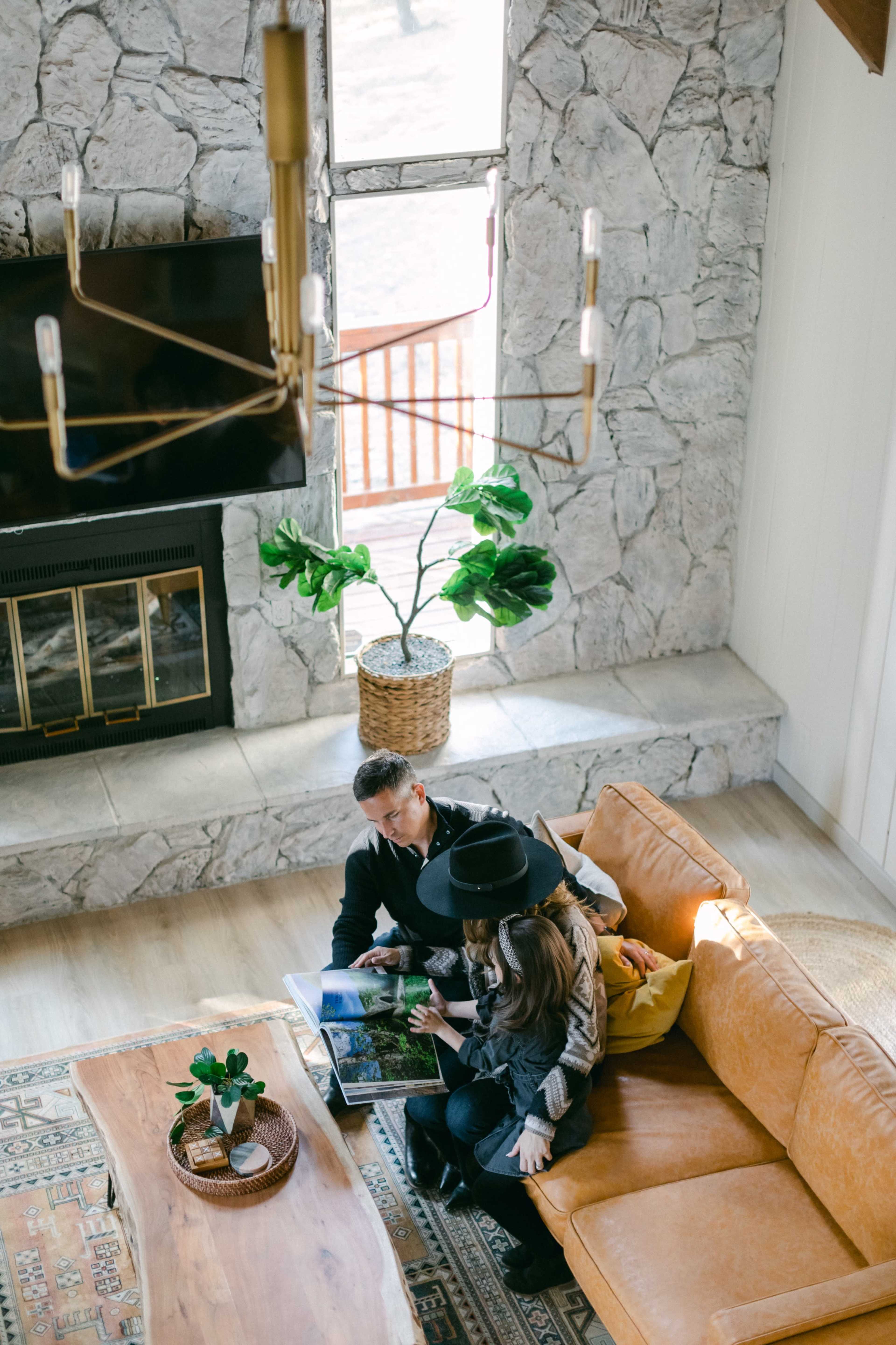 A couple sits together on a brown leather sofa, looking at a photo book in a cozy living room with a stone fireplace and a large window.
