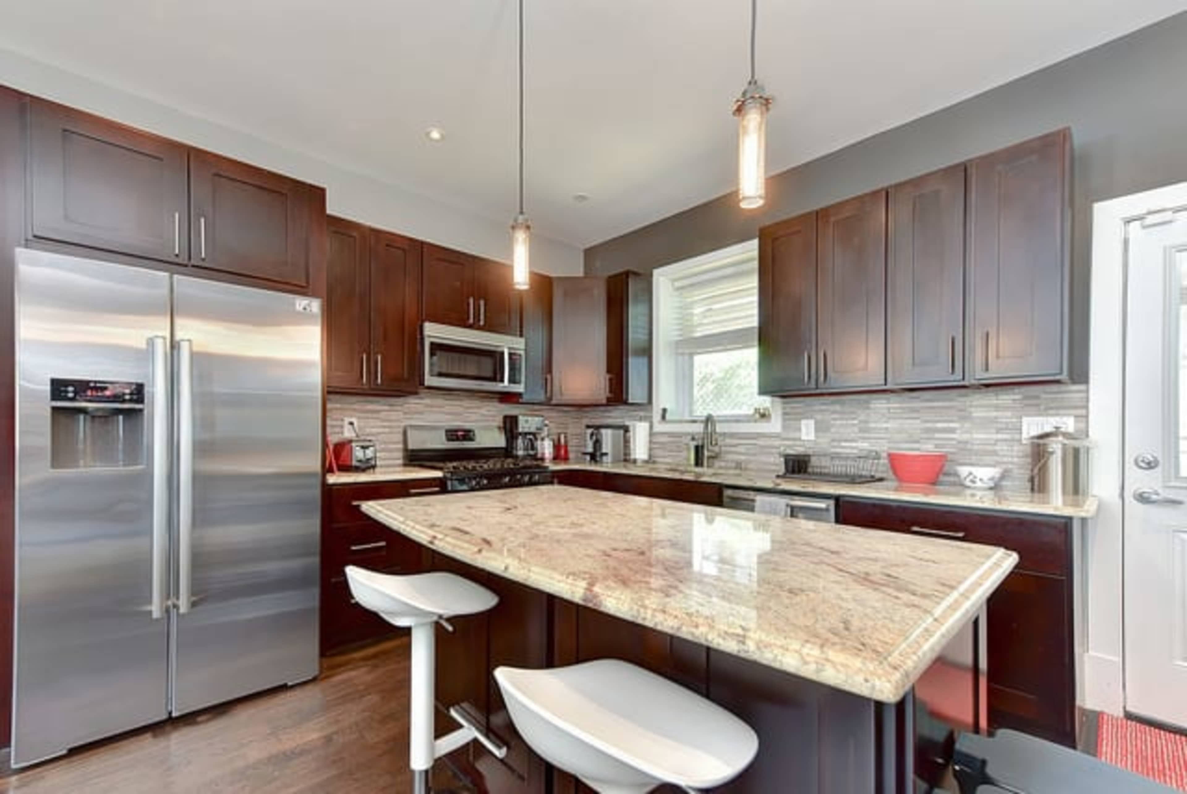 A modern kitchen features dark wooden cabinets, stainless steel appliances, and a large granite island with white bar stools.