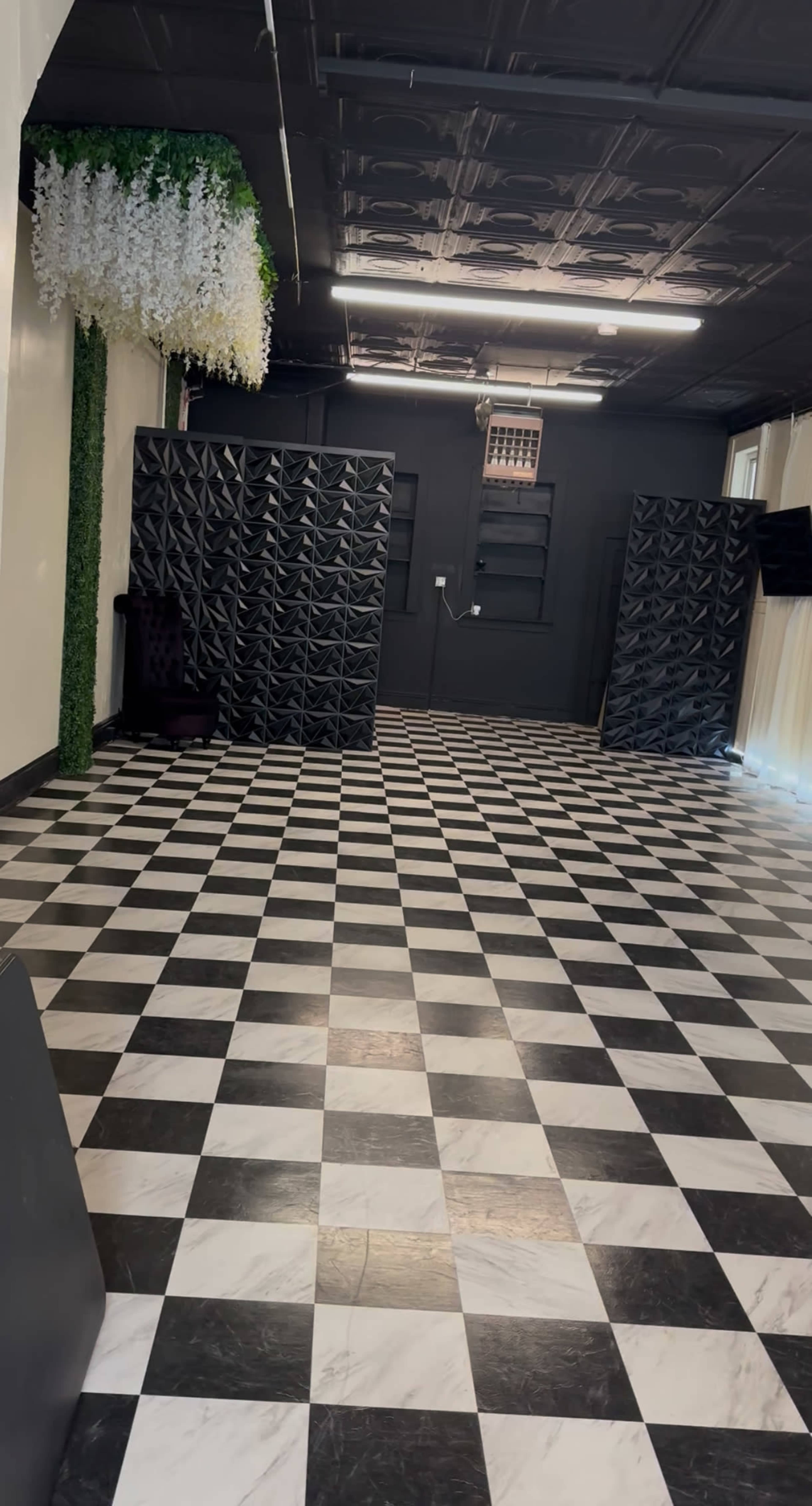 The image shows an empty room with a black and white checkered floor, featuring black dividers on one side and decorative greenery on the ceiling.