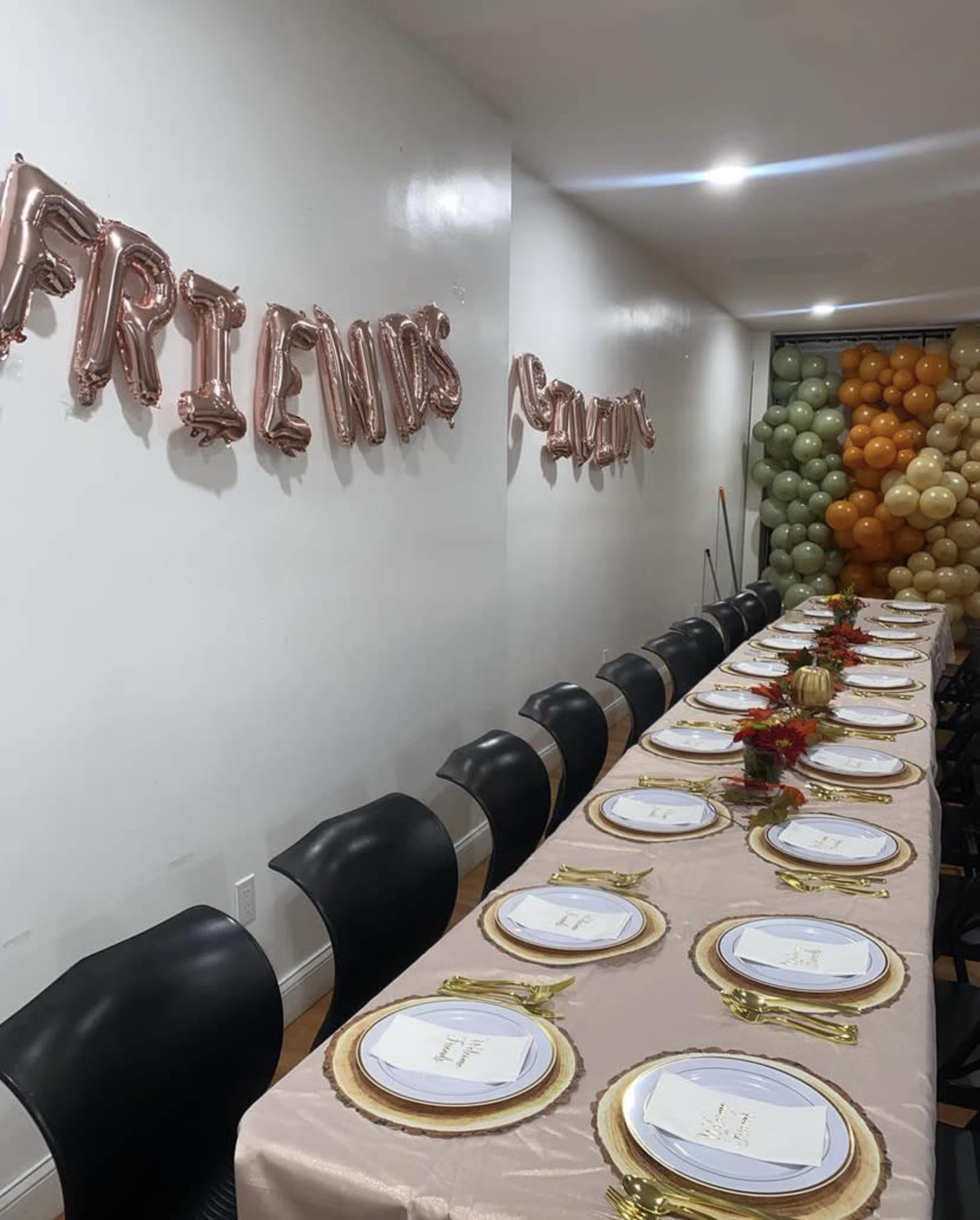 A long dining table is set with elegant plates and golden utensils, surrounded by black chairs, under a "FRIENDS" balloon decoration and a backdrop of colorful balloons.