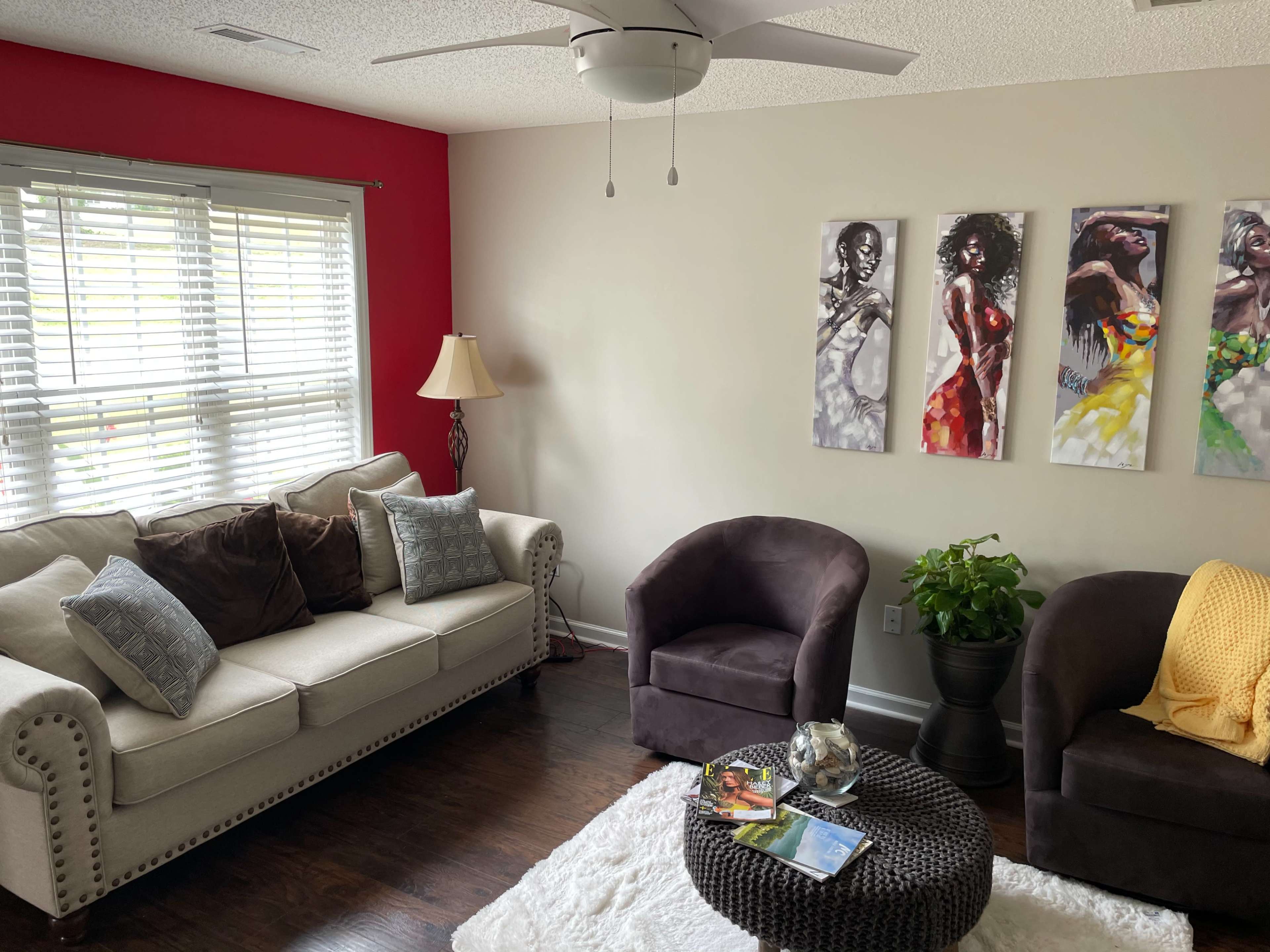 The living room features a light-colored sofa with decorative cushions, two dark armchairs, a round coffee table with books and a plant, and three paintings on the wall, with a red accent wall behind.