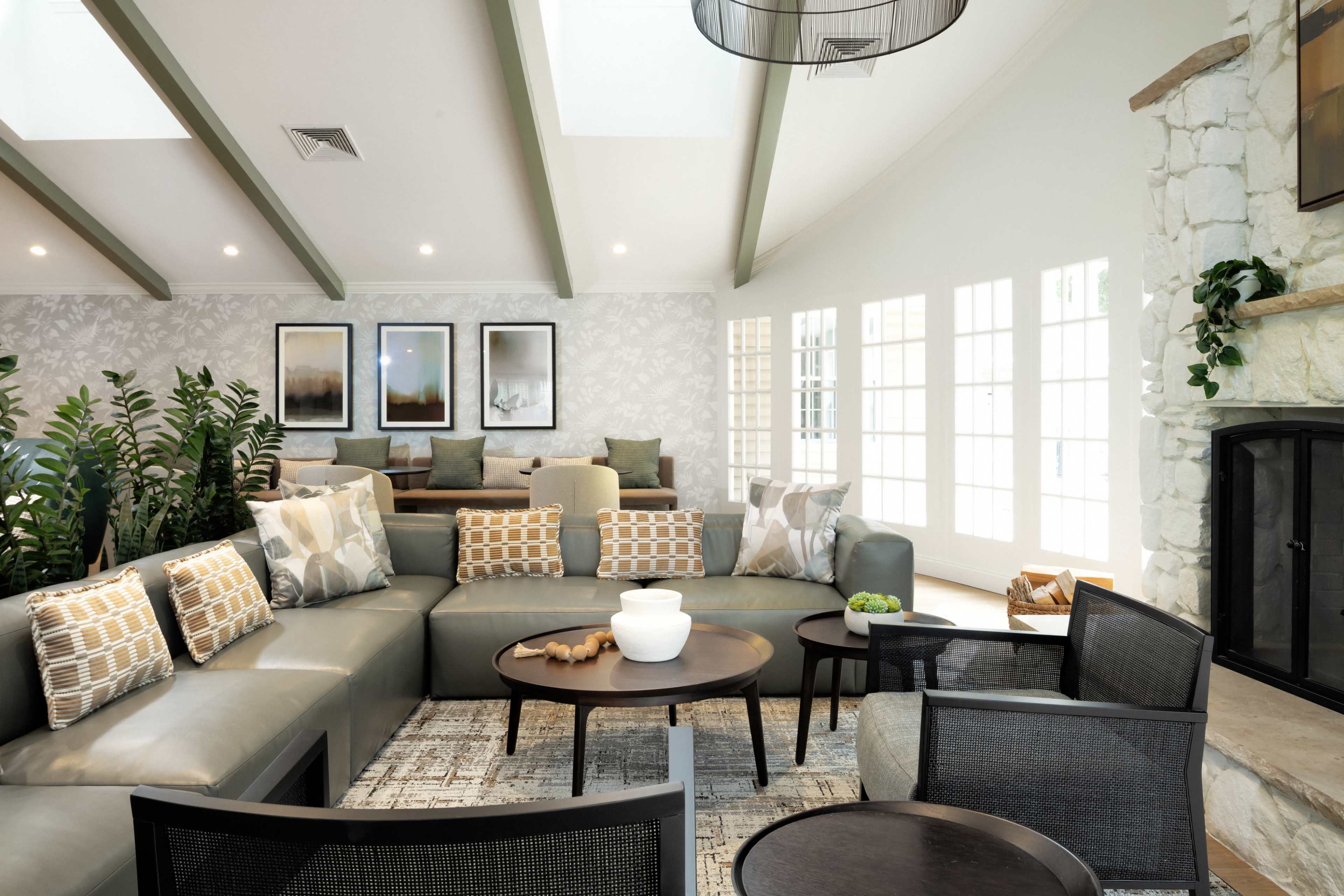 A modern living room features a sectional sofa, a round coffee table, and large windows, with decorative plants and framed artwork on the walls.