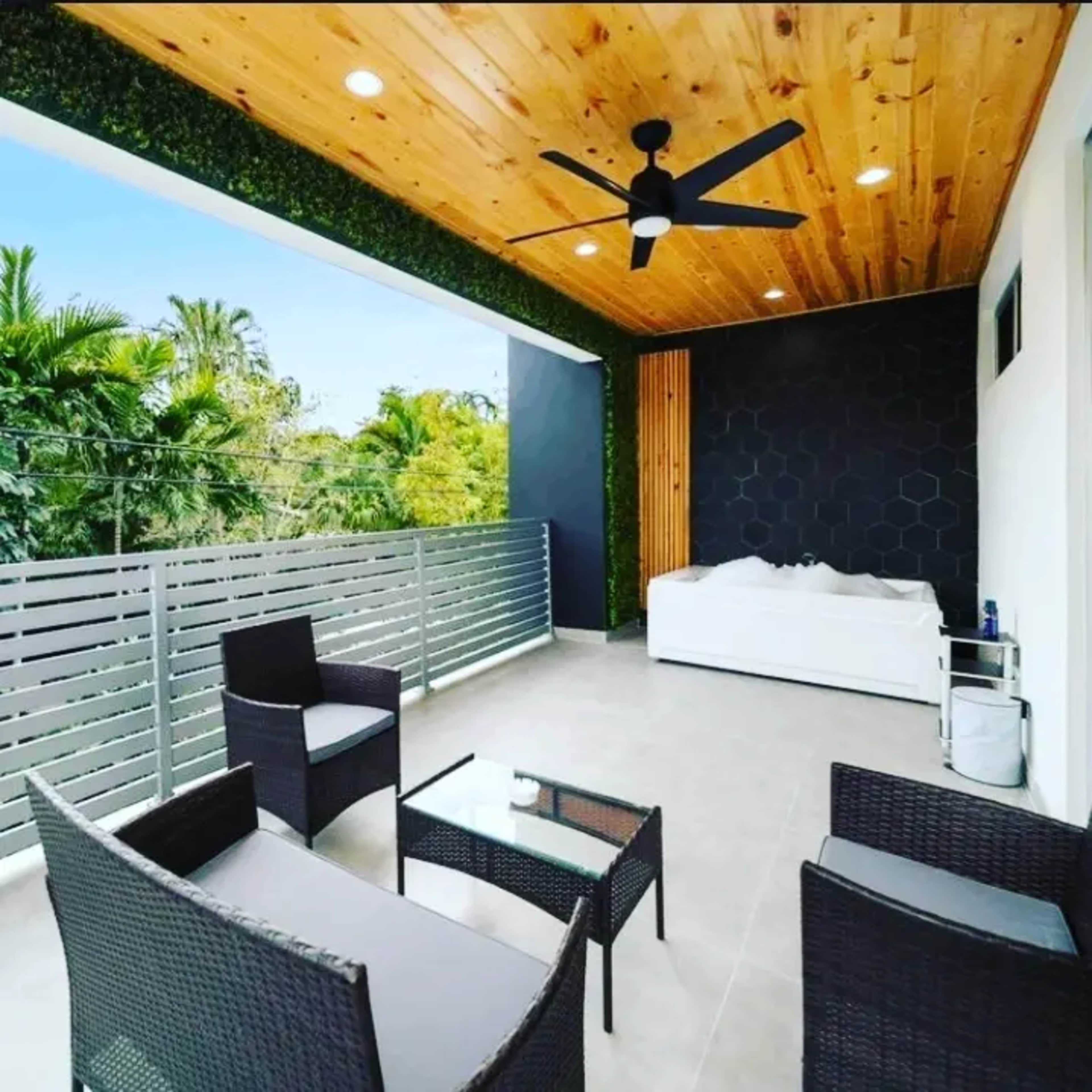 The image depicts a modern outdoor balcony featuring a seating area with wicker chairs and a glass table, surrounded by greenery and a wooden ceiling.