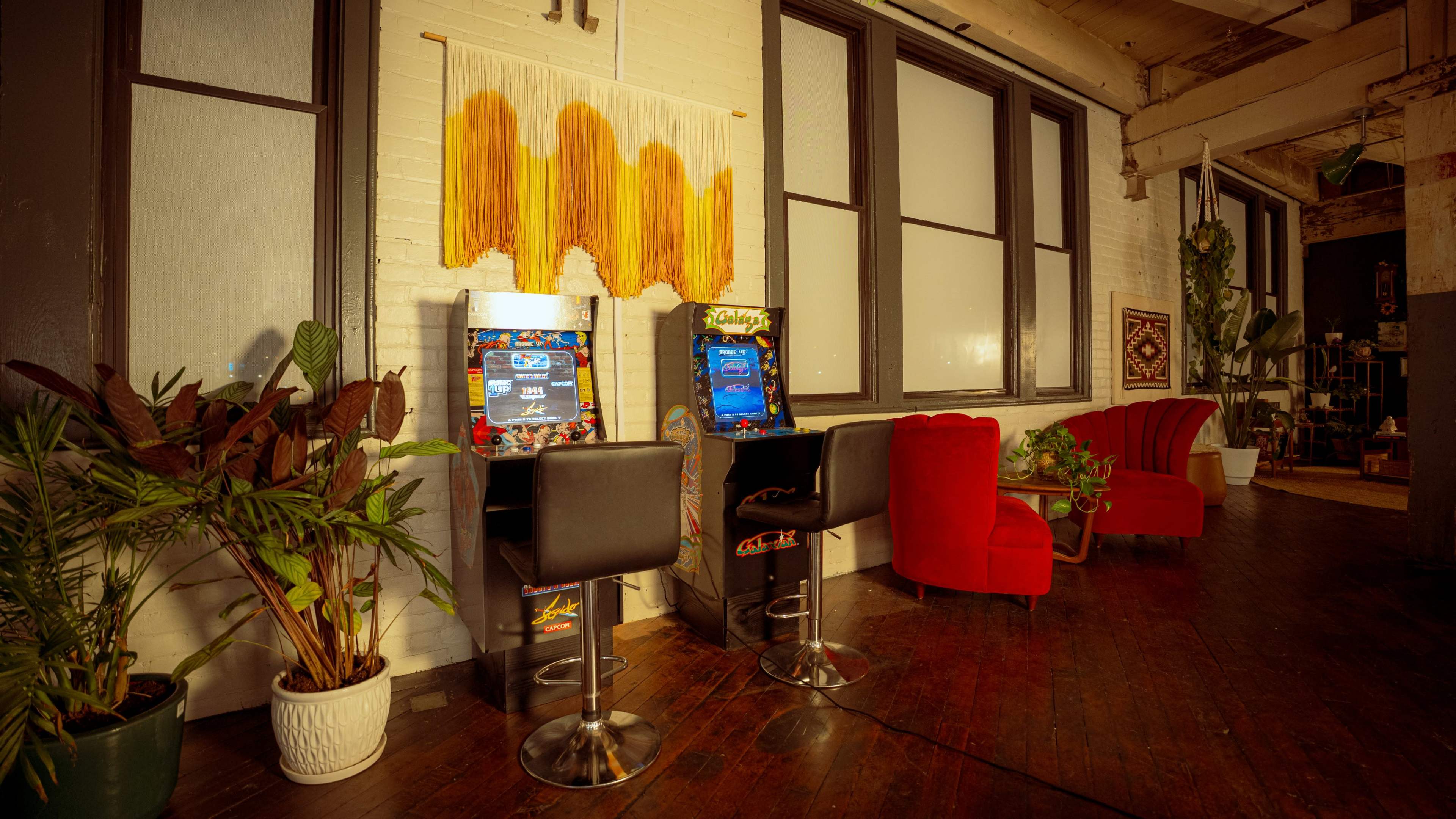 The image shows two vintage arcade game machines placed against a wall in a space with large windows and stylish seating, including red chairs and potted plants.