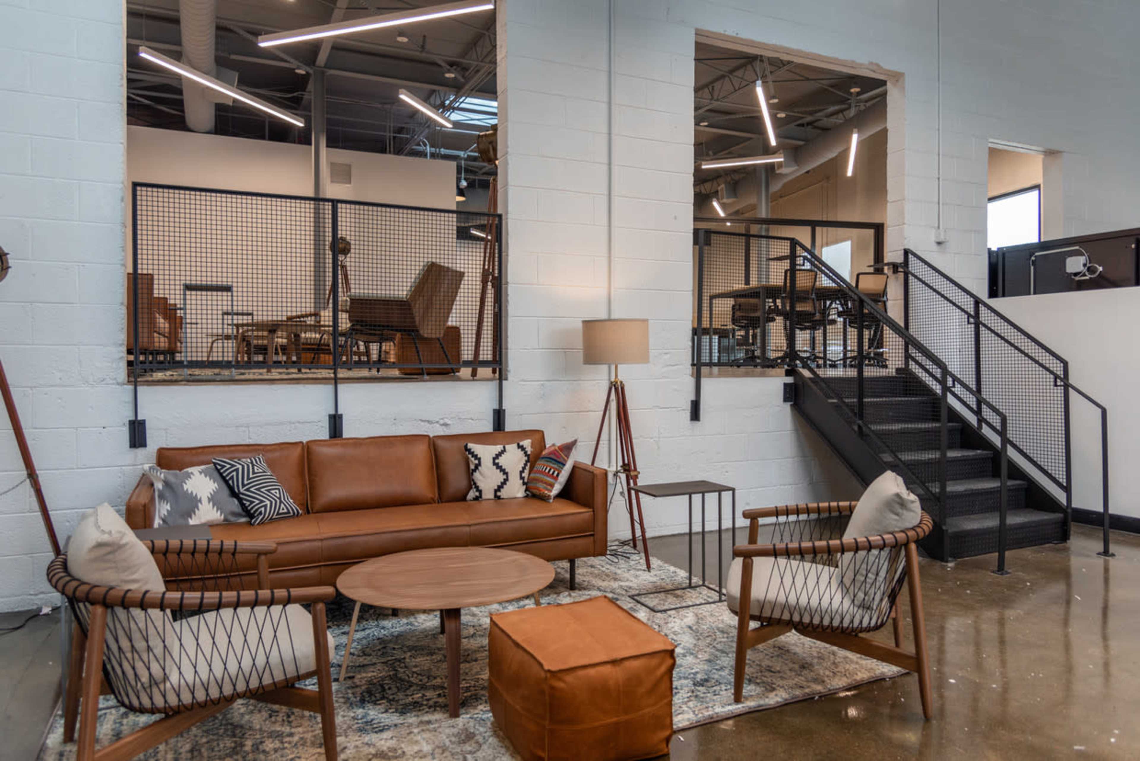 A modern seating area with a brown leather sofa, two woven chairs, a round coffee table, and a lamp, beside a staircase leading to an upper level in a spacious, industrial-style room.