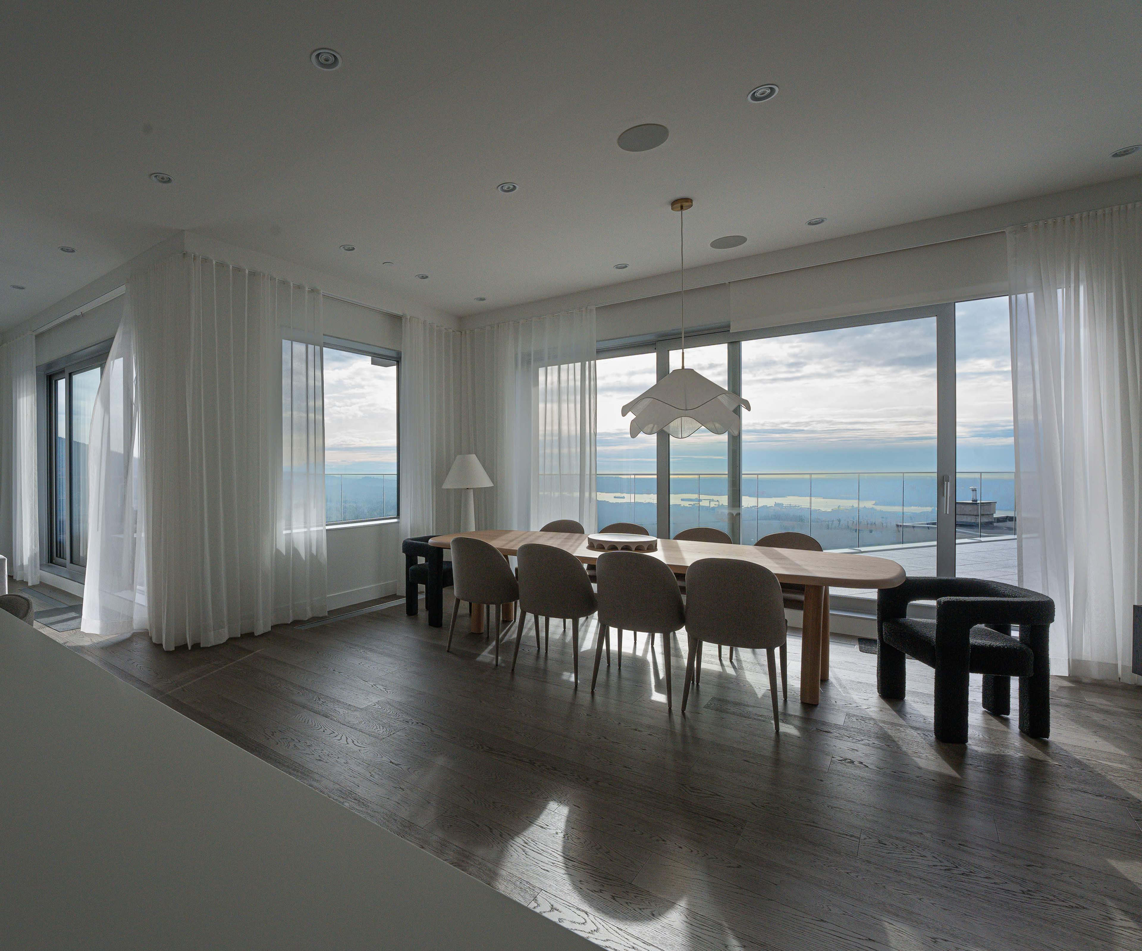 A modern dining area with a long wooden table surrounded by upholstered chairs and large windows that overlook a body of water.