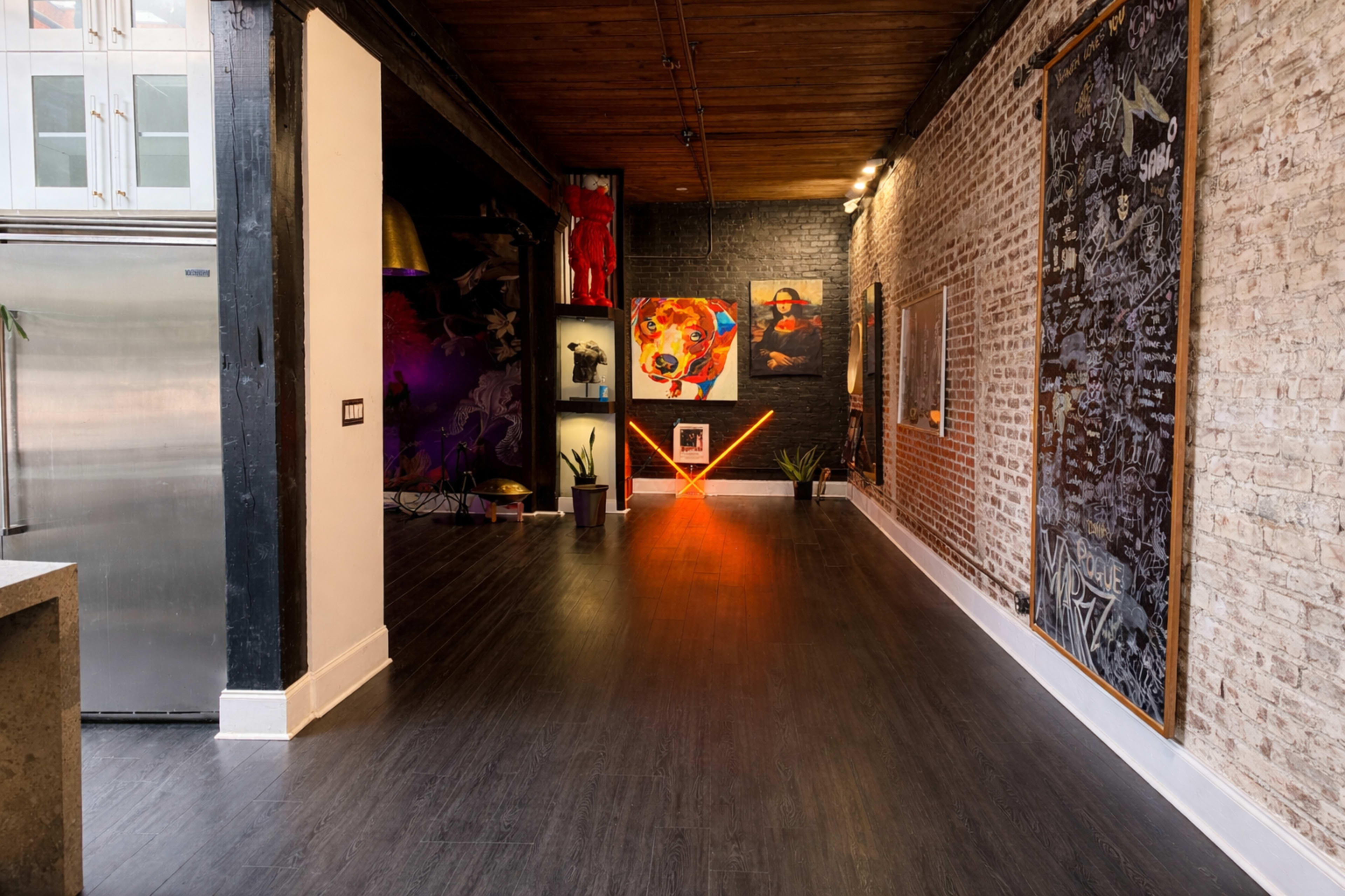 The image shows a spacious art gallery with exposed brick walls, modern lighting, and various colorful artworks displayed on the walls.