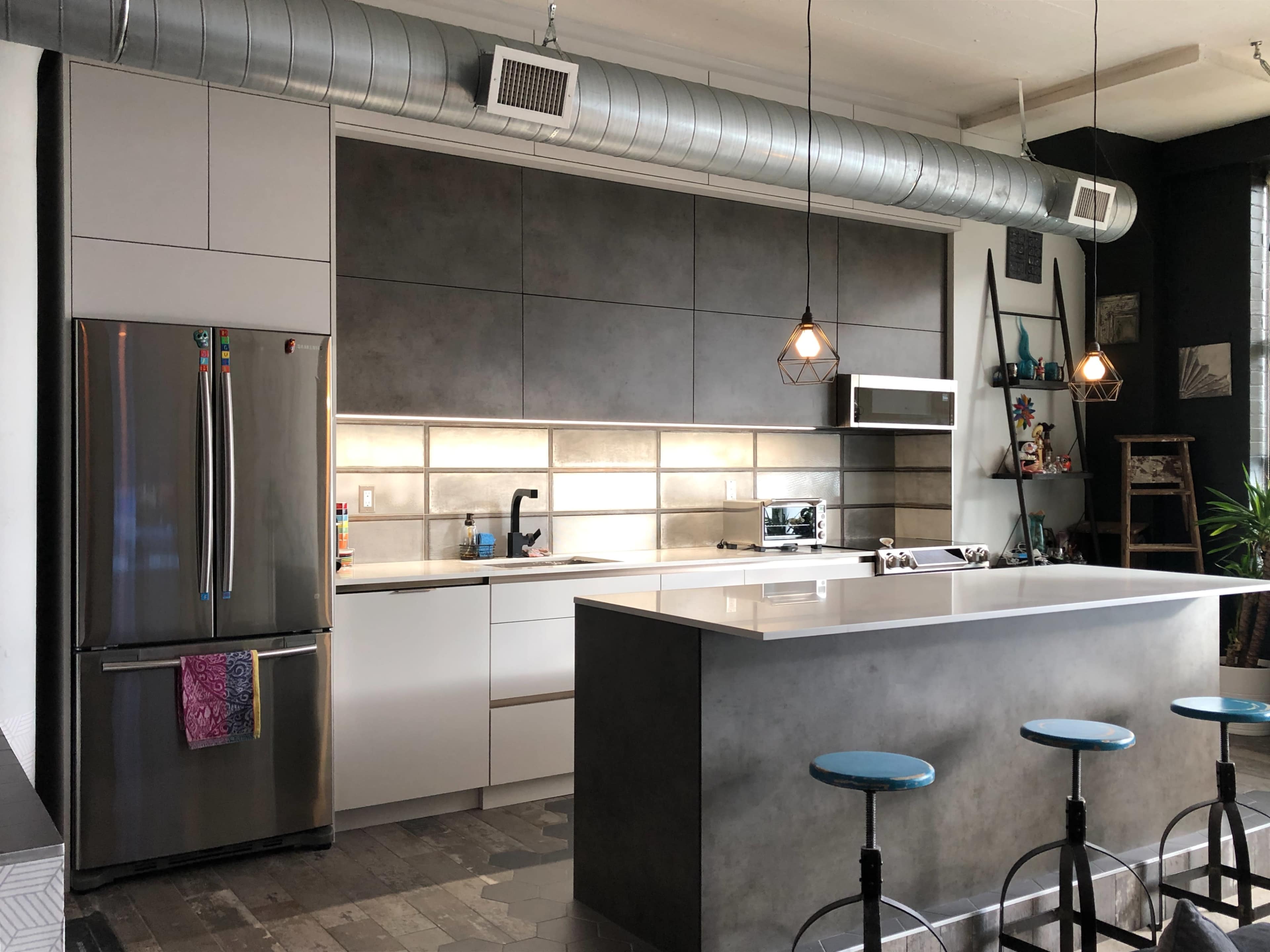 The image shows a modern kitchen with a gray and white color scheme, featuring stainless steel appliances, a large island with blue bar stools, and exposed ductwork.