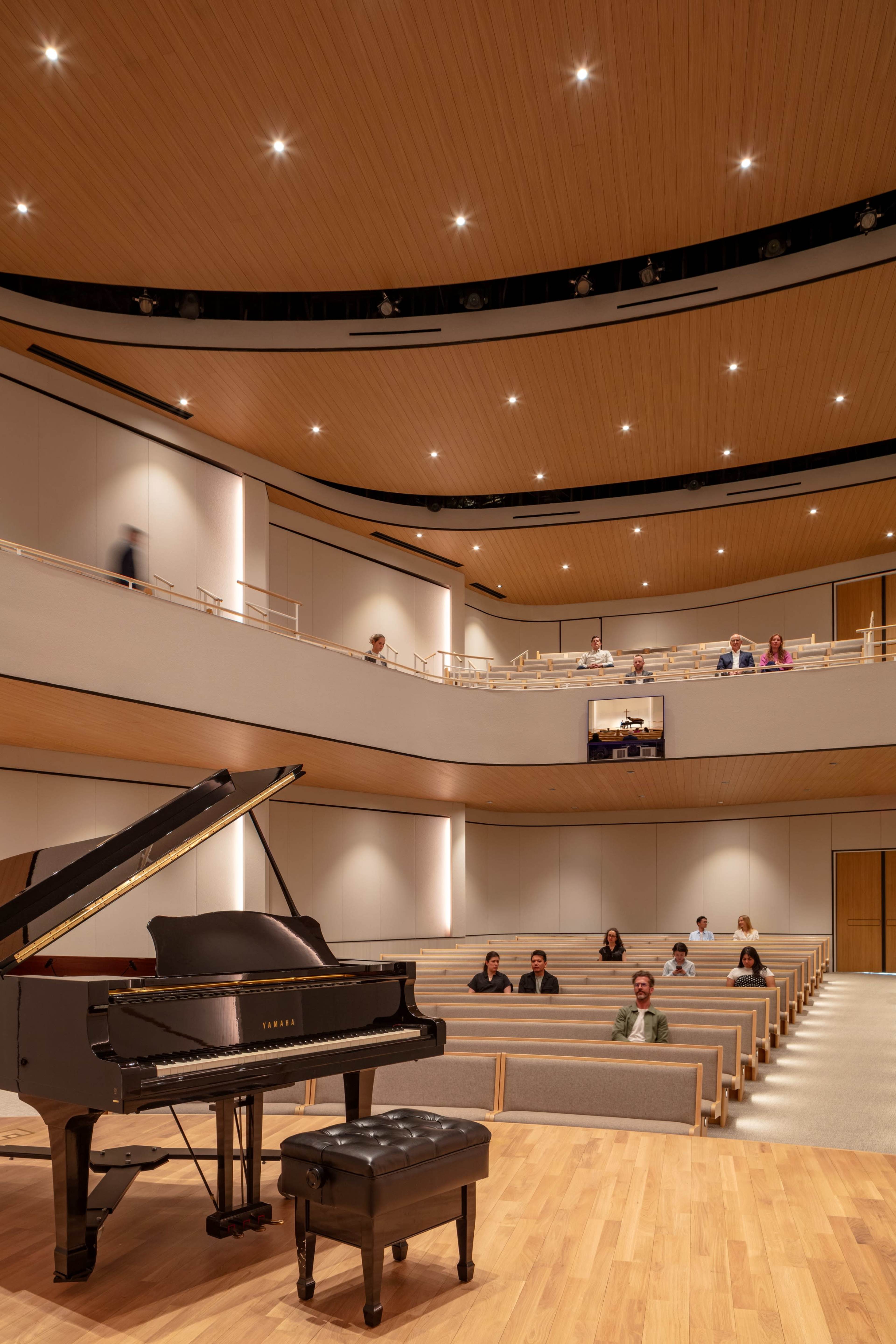 A grand piano sits on a wooden stage in a modern auditorium with tiered seating, where a small audience is attentively watching.