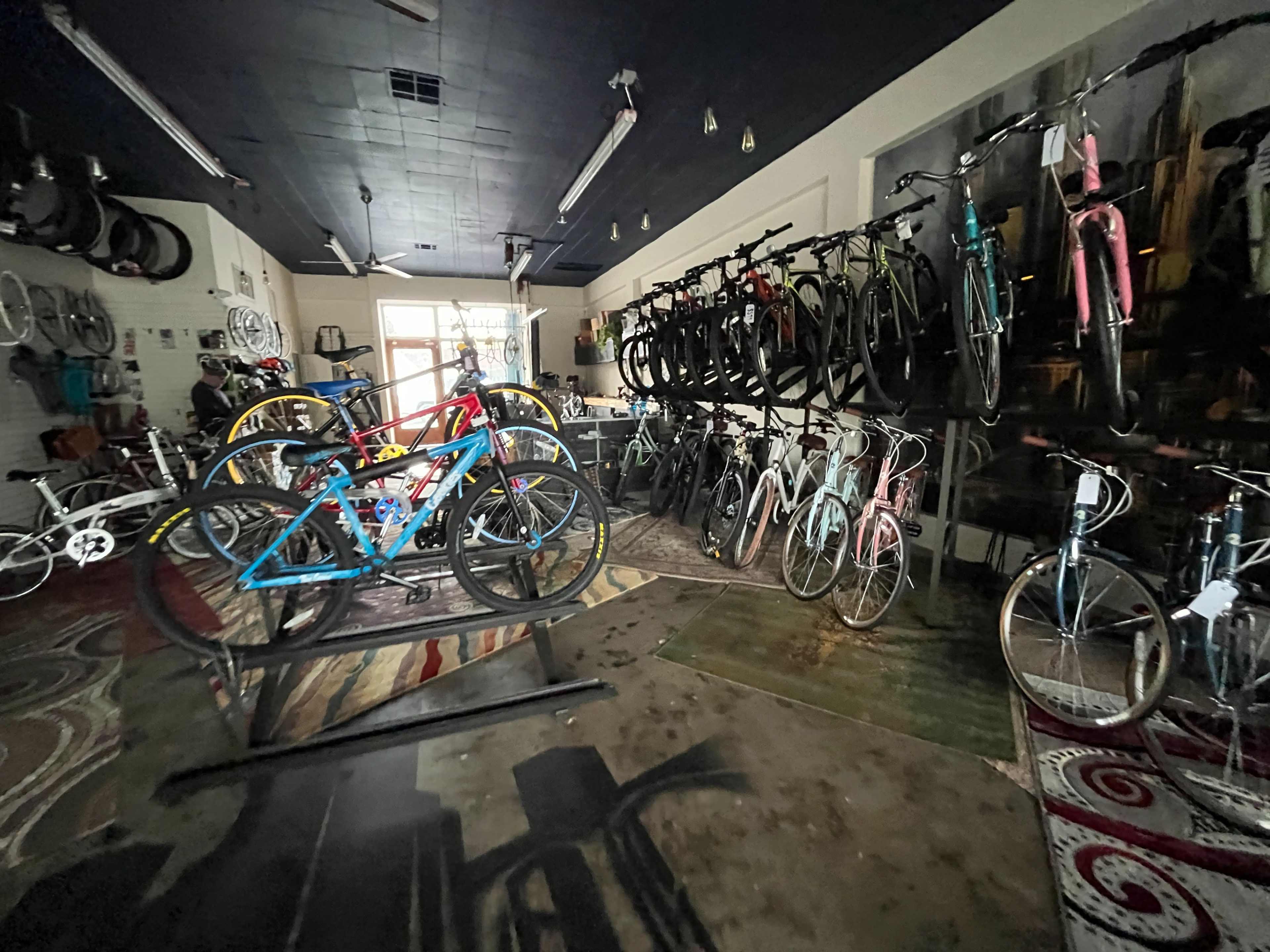 The image shows a bicycle shop with various bikes on display, including colorful mountain and road bikes, arranged on racks and shelves in a cluttered interior.