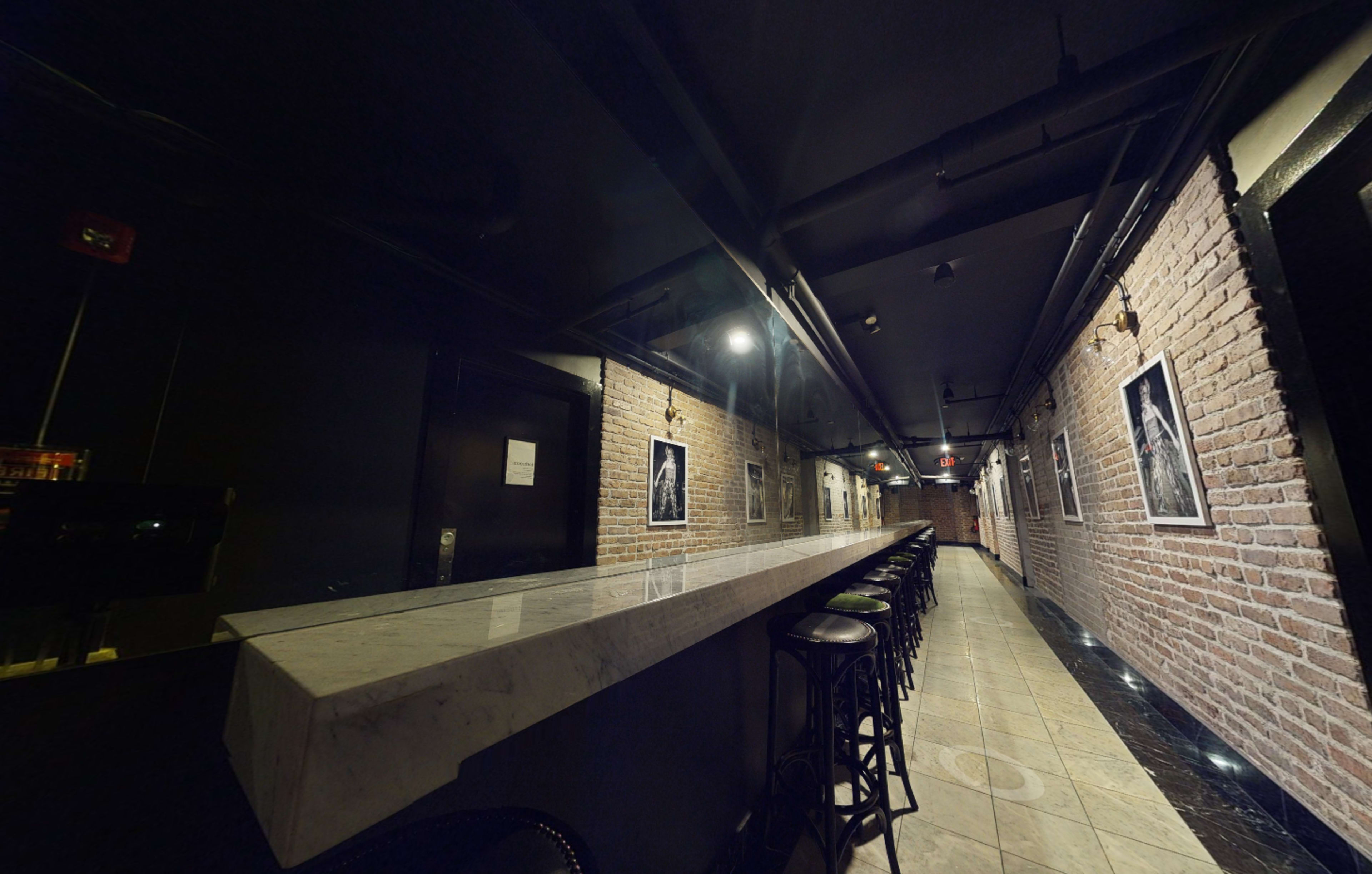 The image shows a long, narrow bar area with a marble counter and black stools, flanked by exposed brick walls and framed photographs.