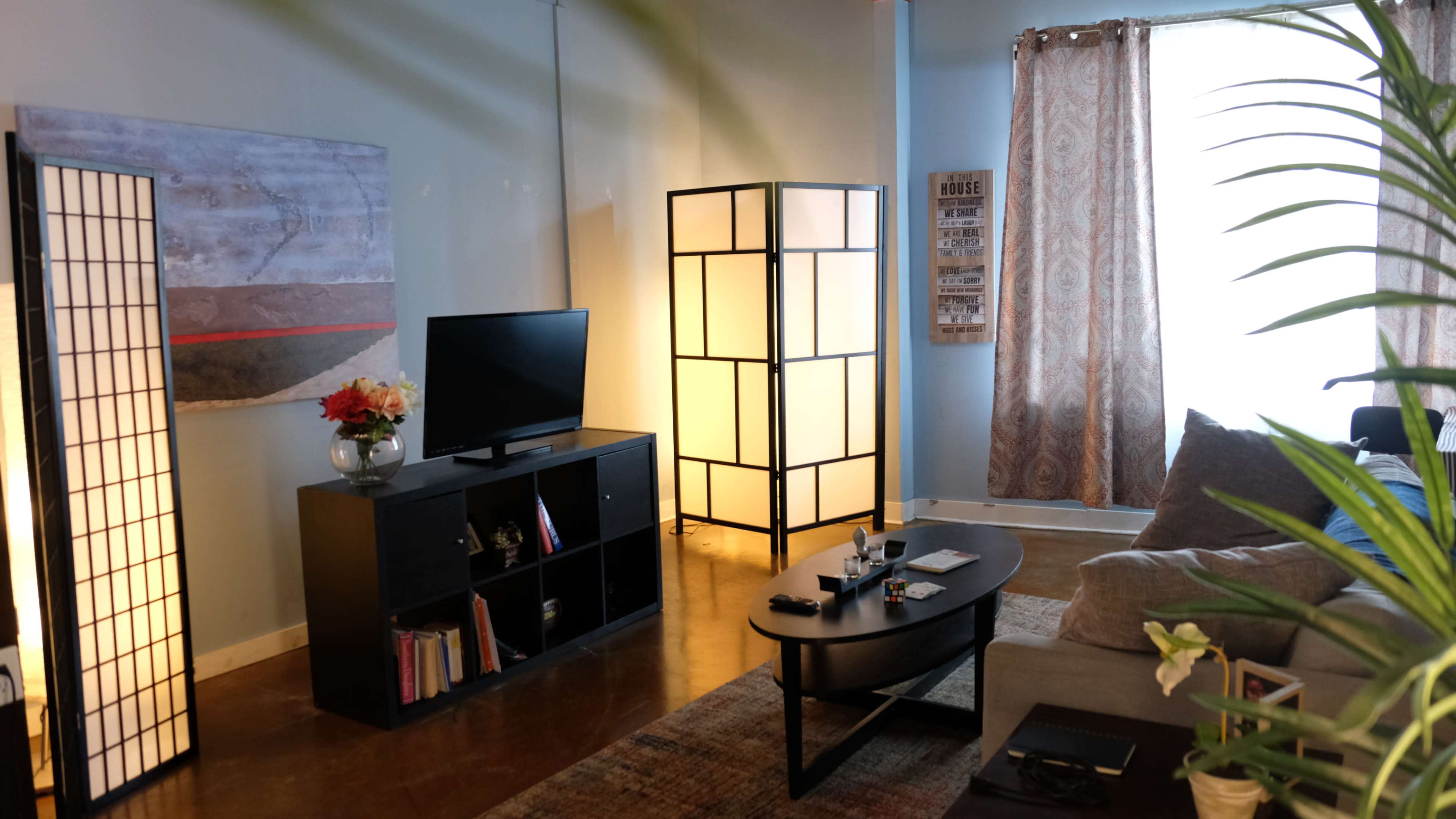 The image shows a living room with a television on a black cabinet, a sofa, a coffee table, and decorative screens.