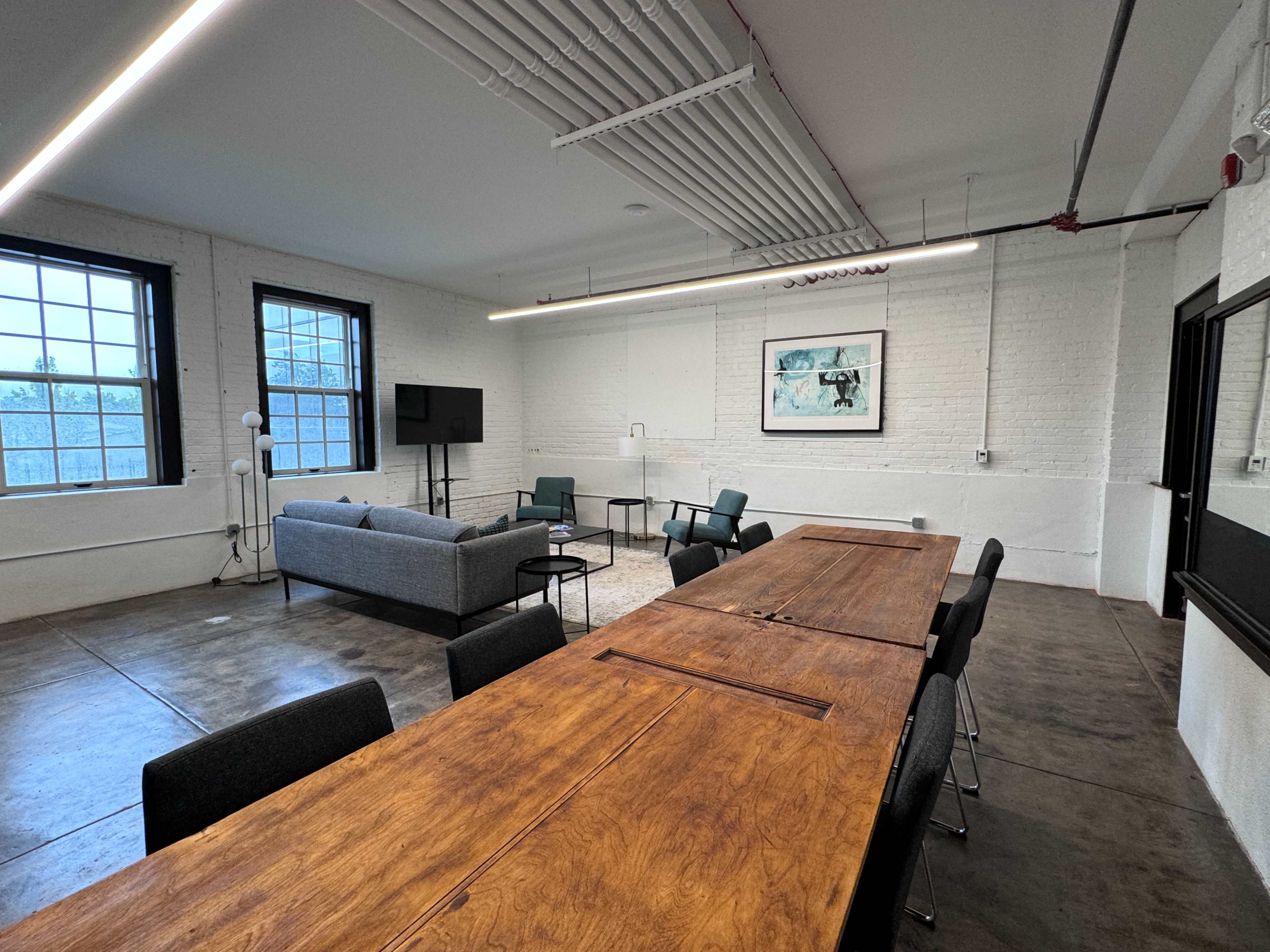 The image shows a modern office space with a long wooden table, a gray sofa, chairs, and large windows letting in natural light.