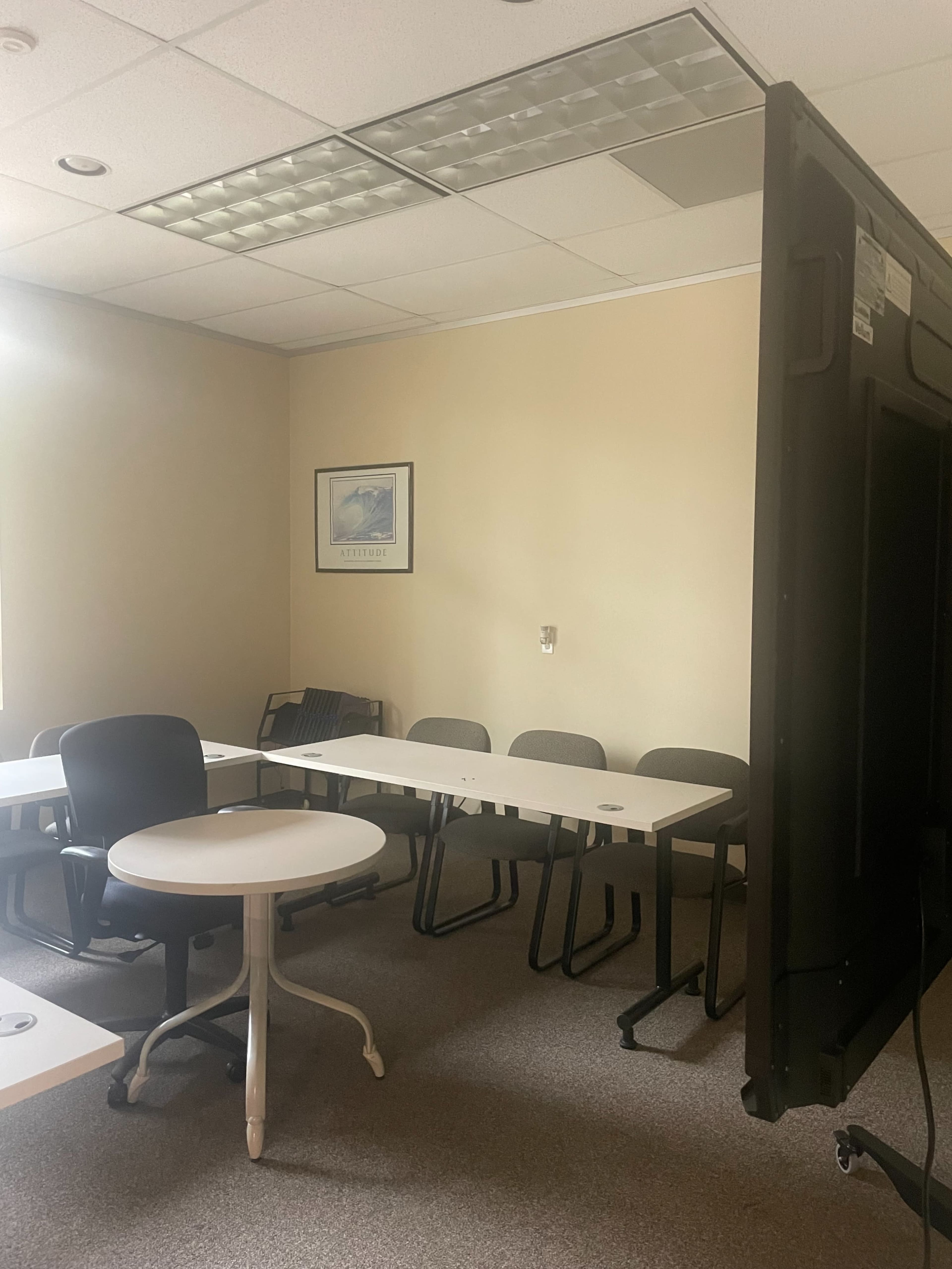 A small meeting room features several tables and chairs arranged, with a large screen positioned against one wall.