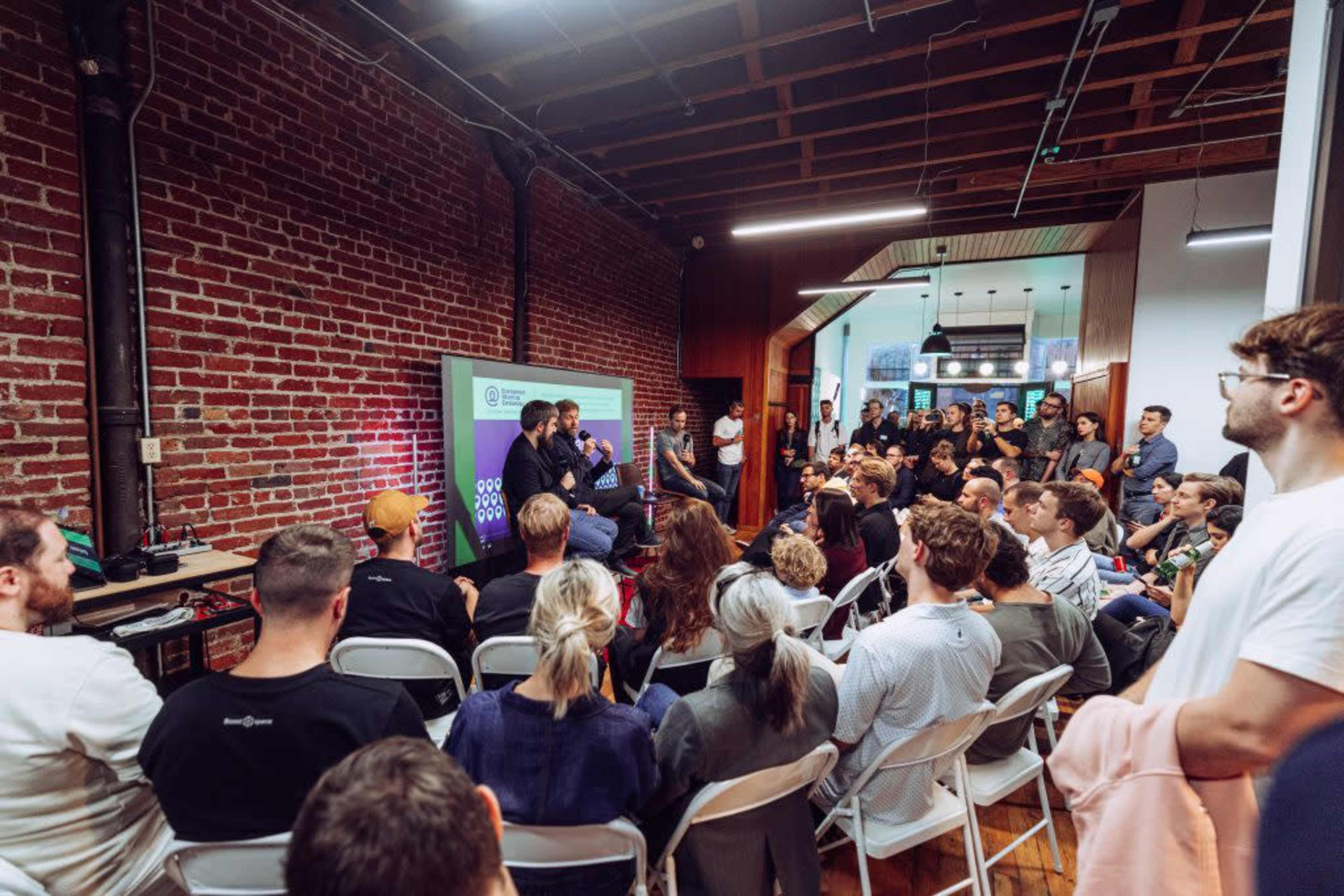 A large audience sits in a room with exposed brick walls, listening to speakers on a stage during an event.