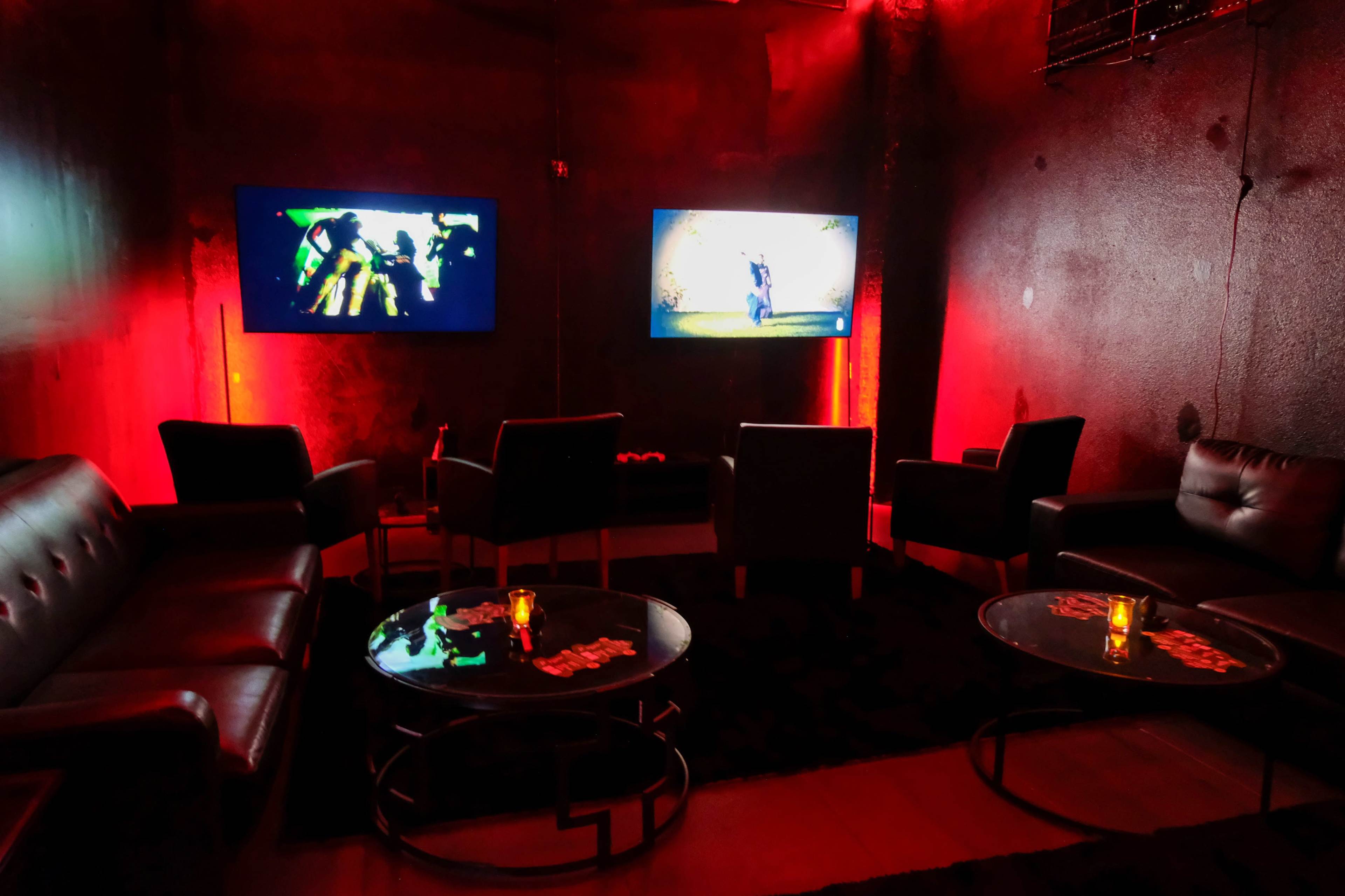 The room features two large screens displaying images, surrounded by black leather sofas and low tables with candles, all illuminated by red lighting.
