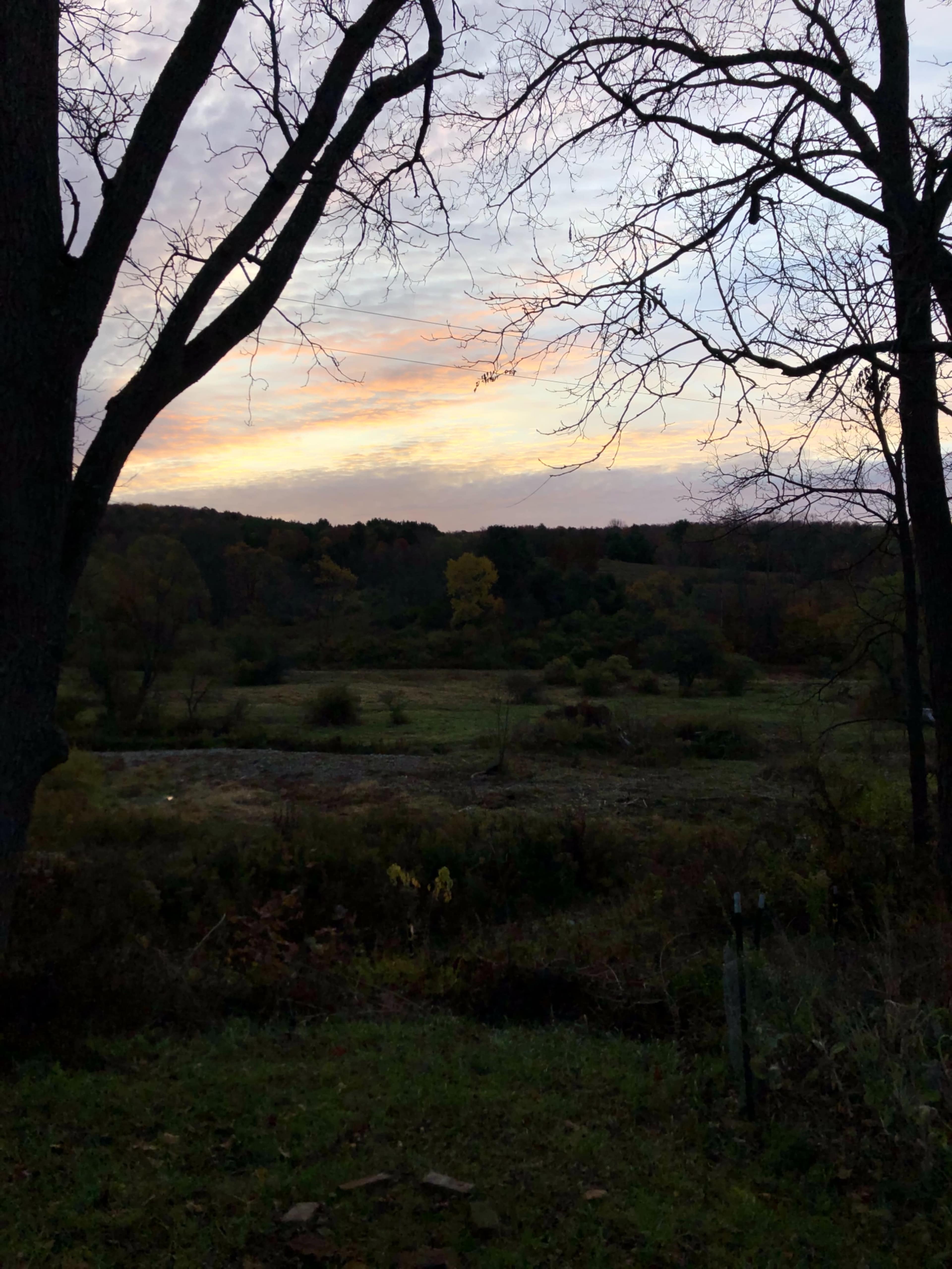 The image shows a sunset over a rural landscape framed by bare tree branches.