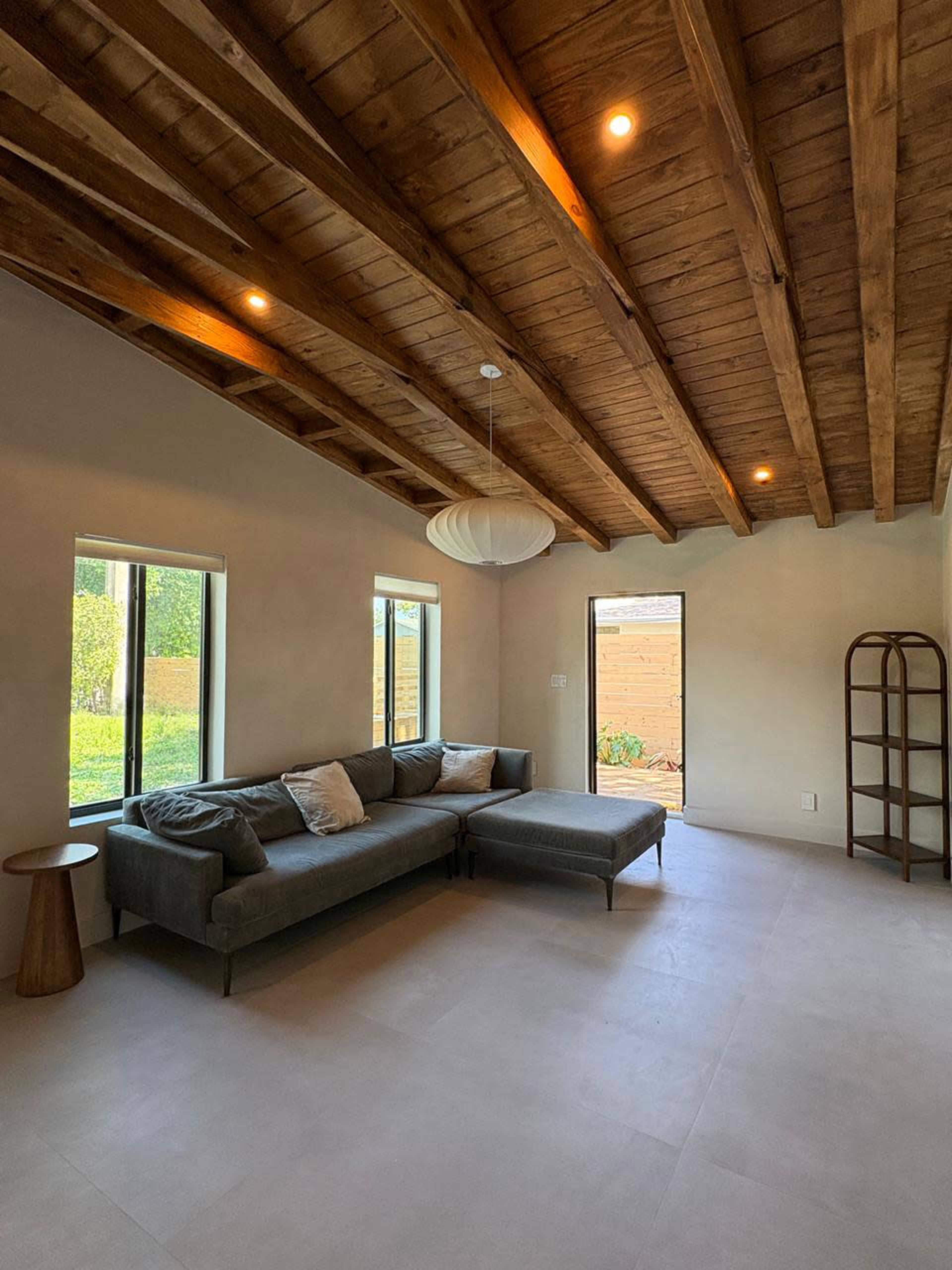 A living room features a gray sectional sofa, a wooden coffee table, large windows, and a pendant light, with exposed wooden beams on the ceiling.