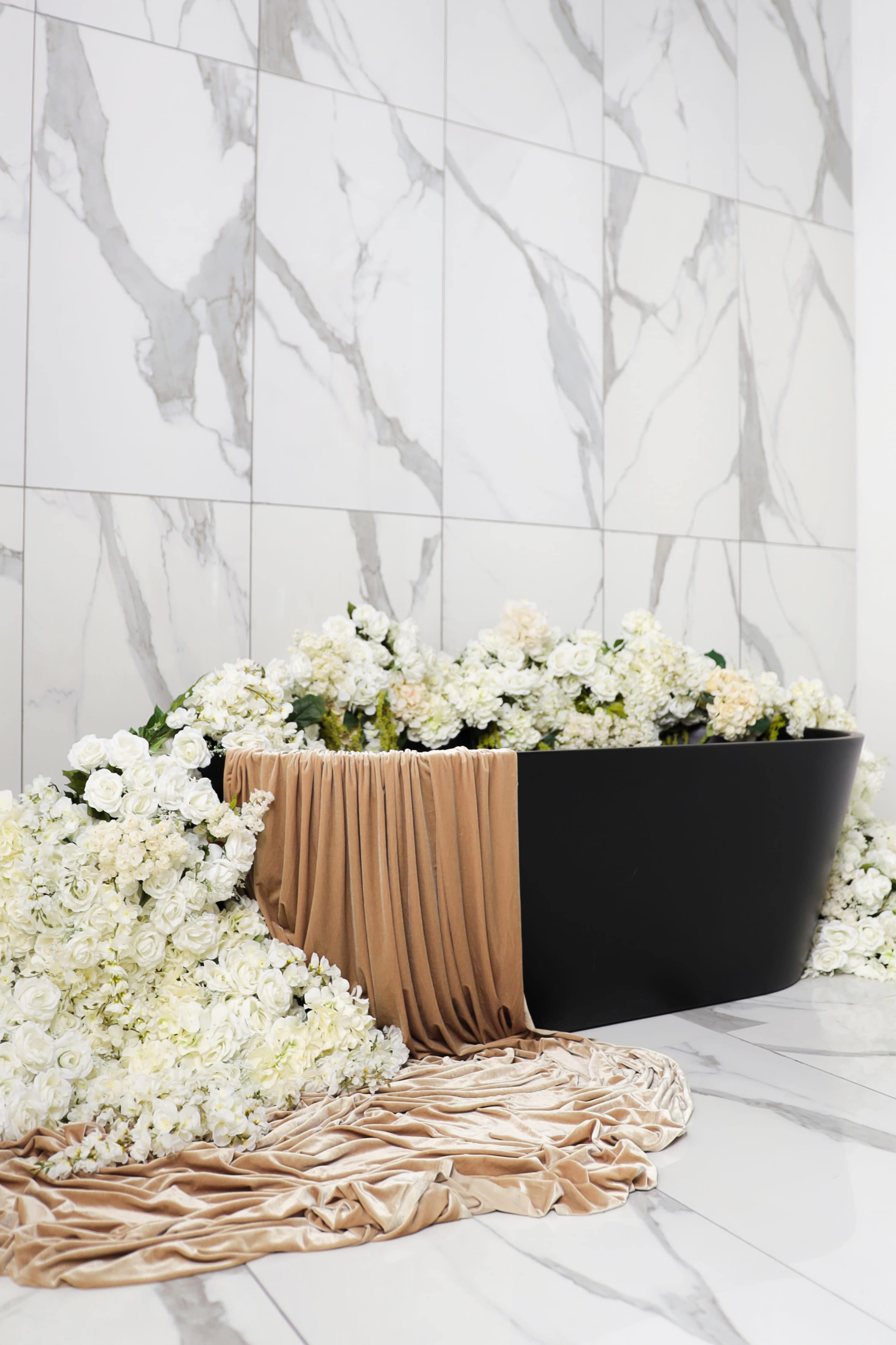 A black bathtub is surrounded by an abundance of white flowers and draped fabric on a marble floor.