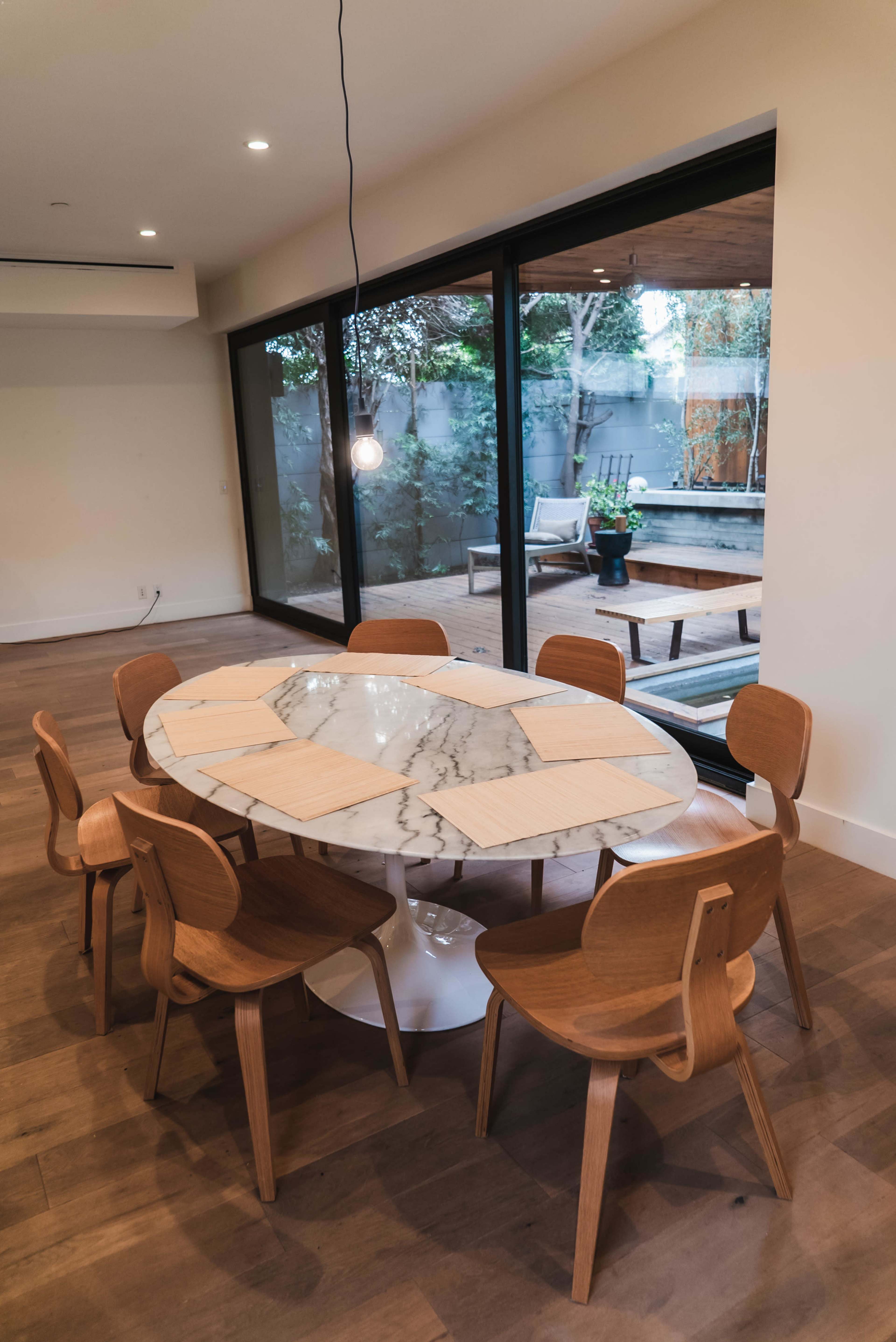 A round marble dining table with six wooden chairs is positioned in a room that features large glass doors opening to a patio view.