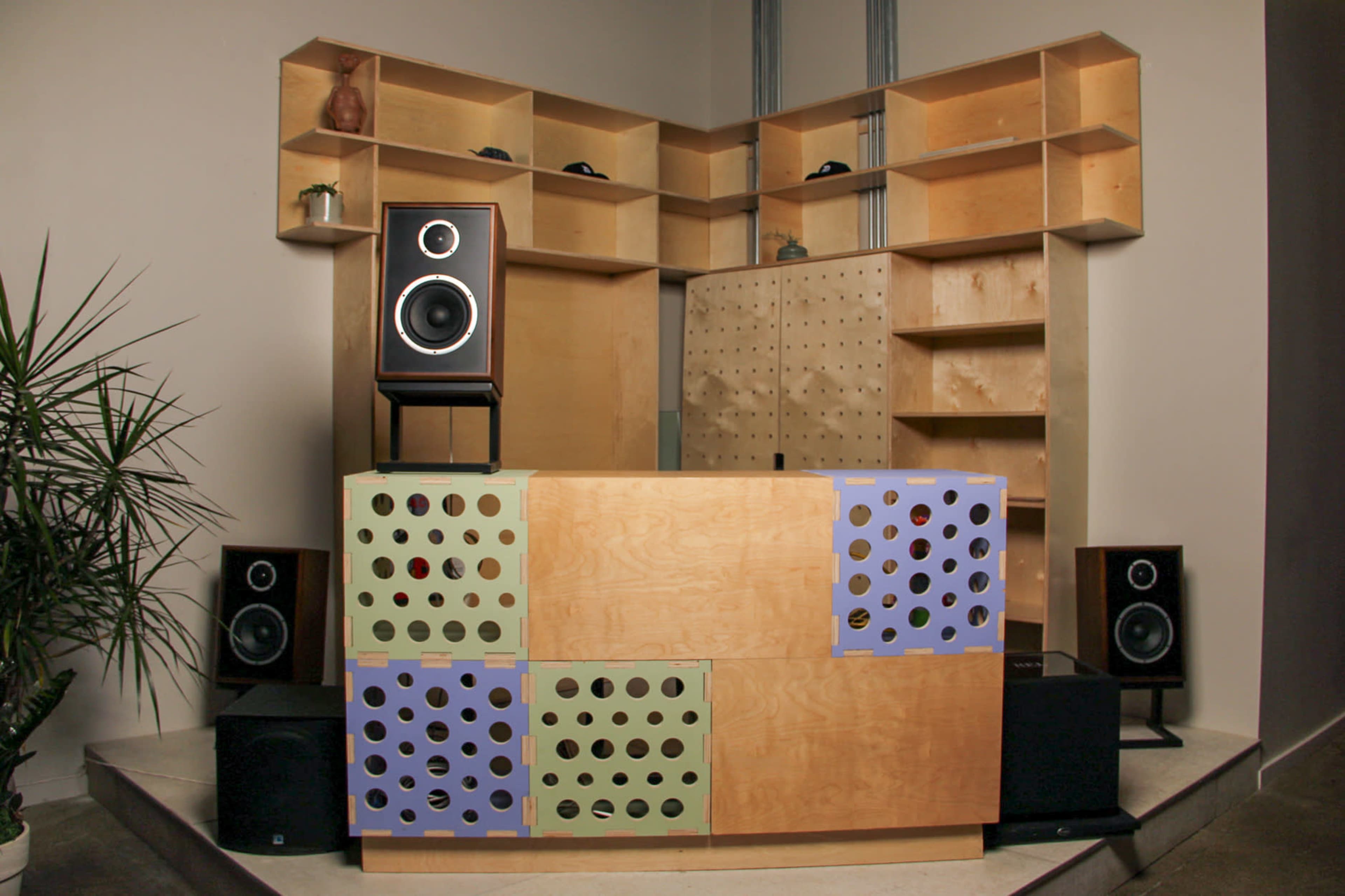 The image shows a modern reception or DJ booth made of wood, featuring a unique design with colorful perforated panels, surrounded by speakers and shelving.