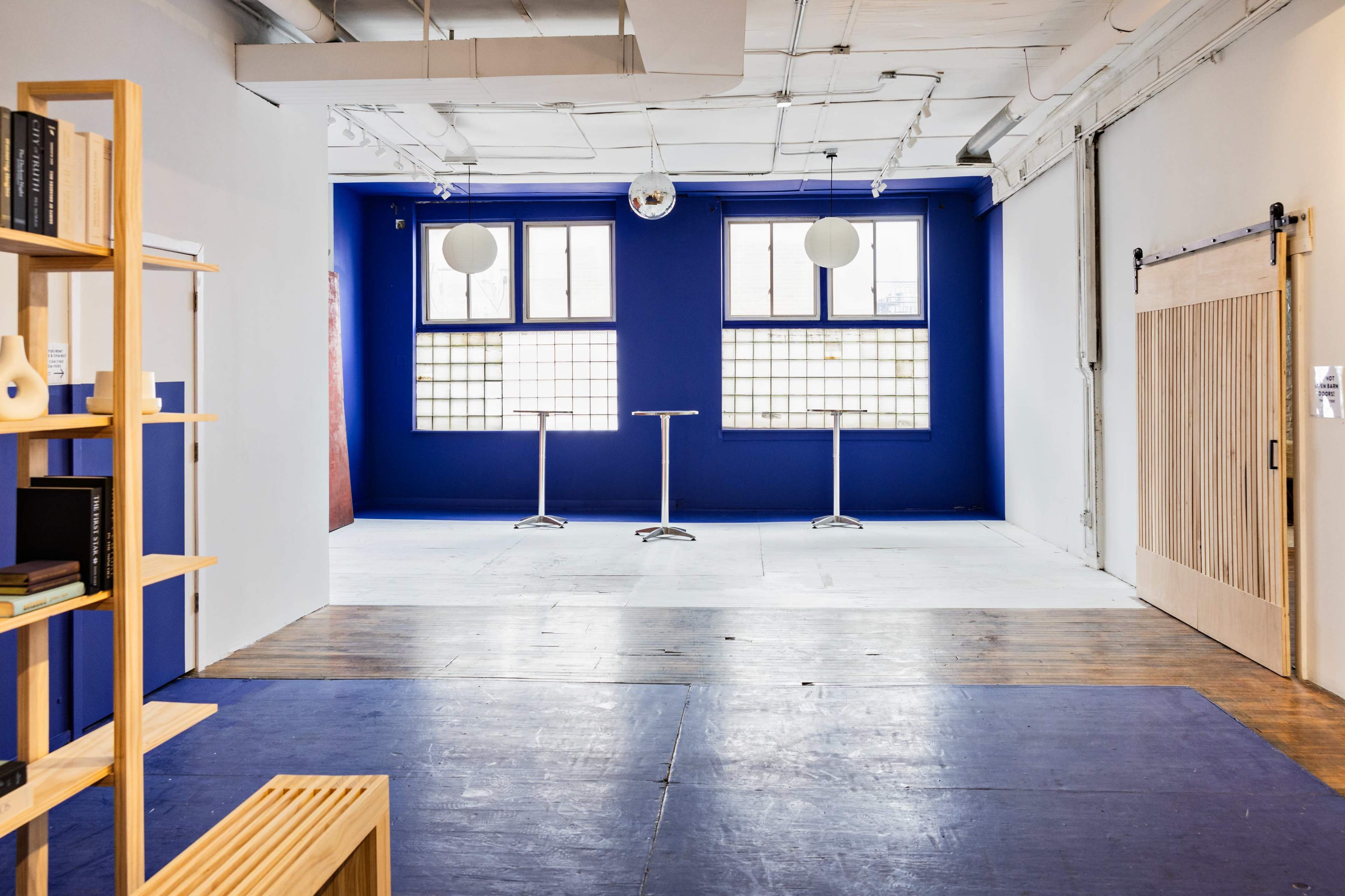 The image shows a spacious, well-lit interior with blue walls, two tall tables near large windows, and a wooden sliding door on the right.