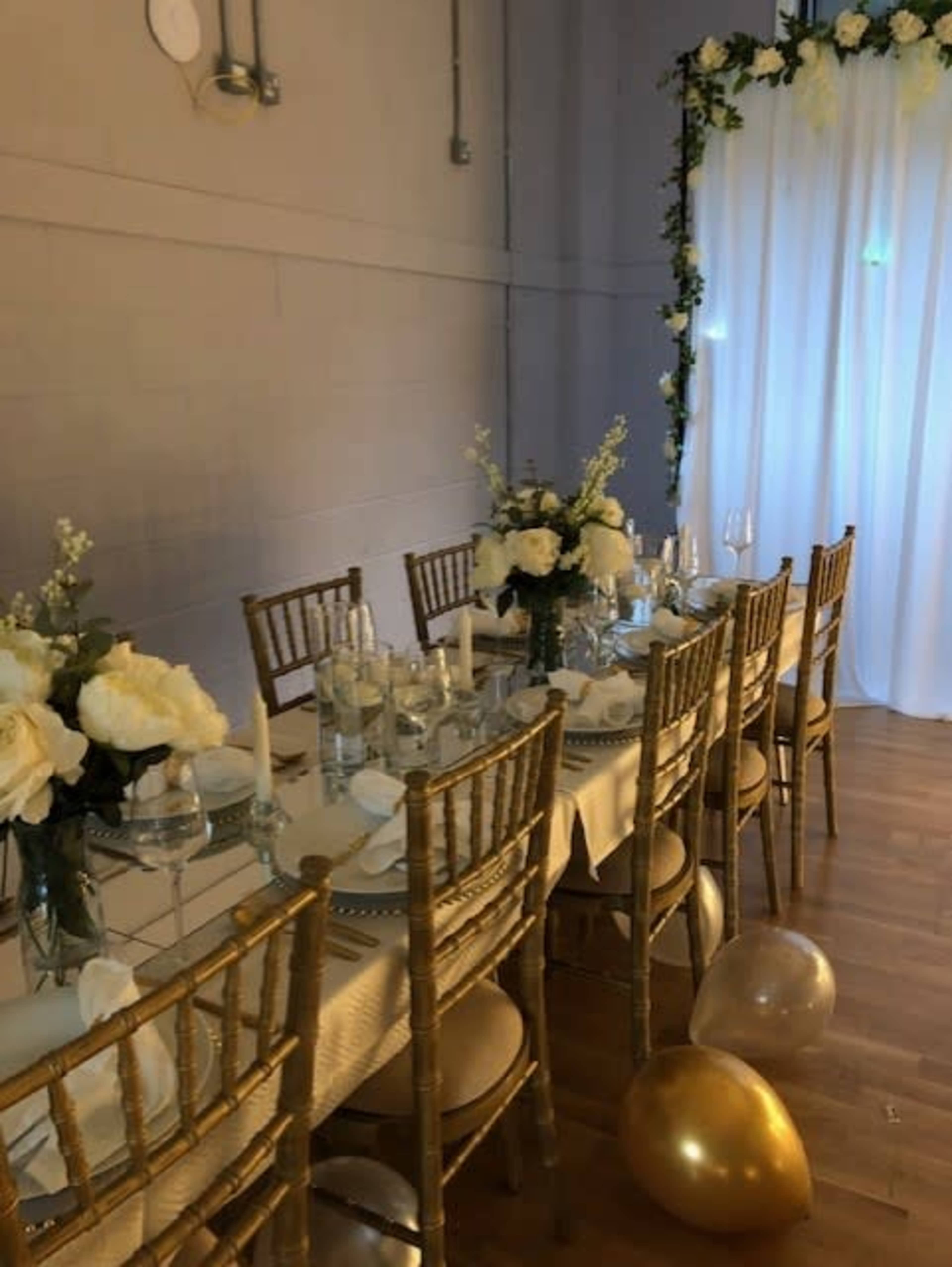 A long dining table is set with elegant place settings, floral arrangements, and golden chairs in a softly lit room.
