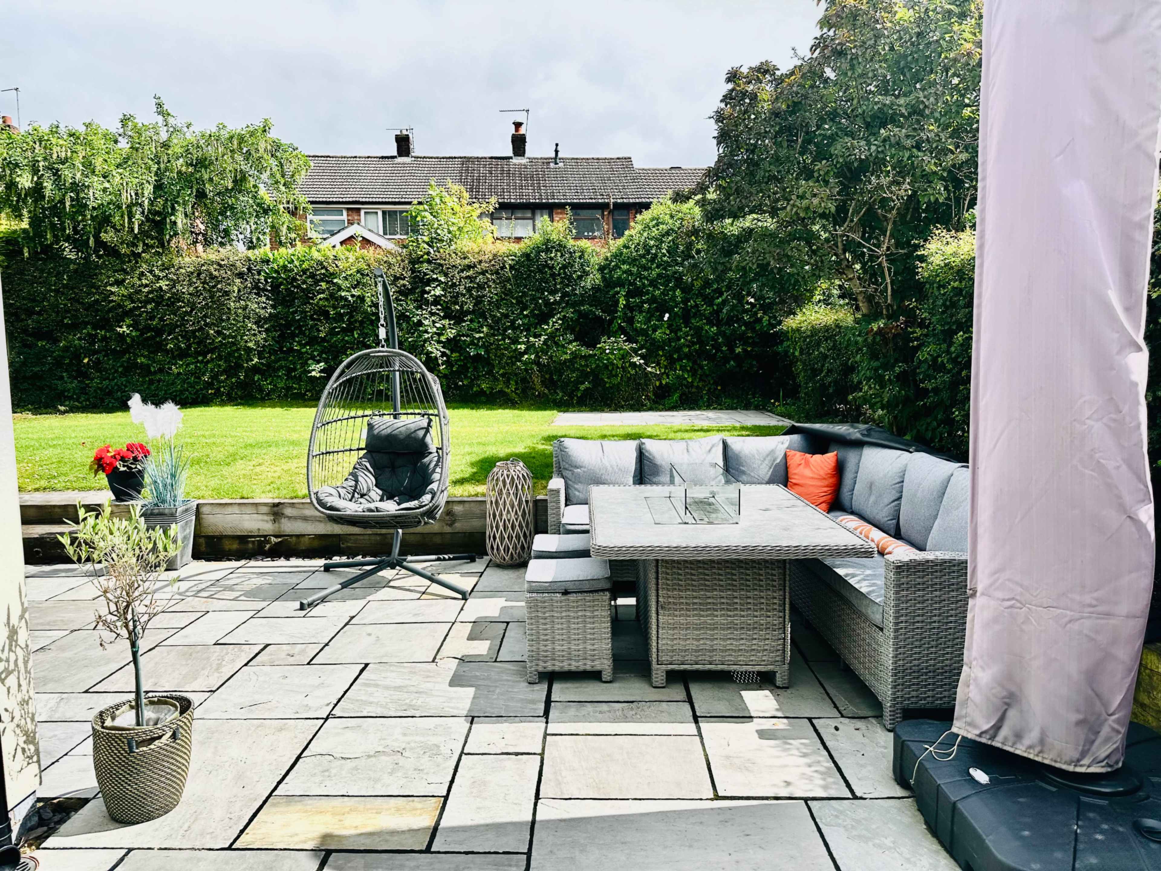 The image shows a landscaped backyard with a stone patio, a corner seating area with a glass table, and a hanging chair surrounded by greenery.