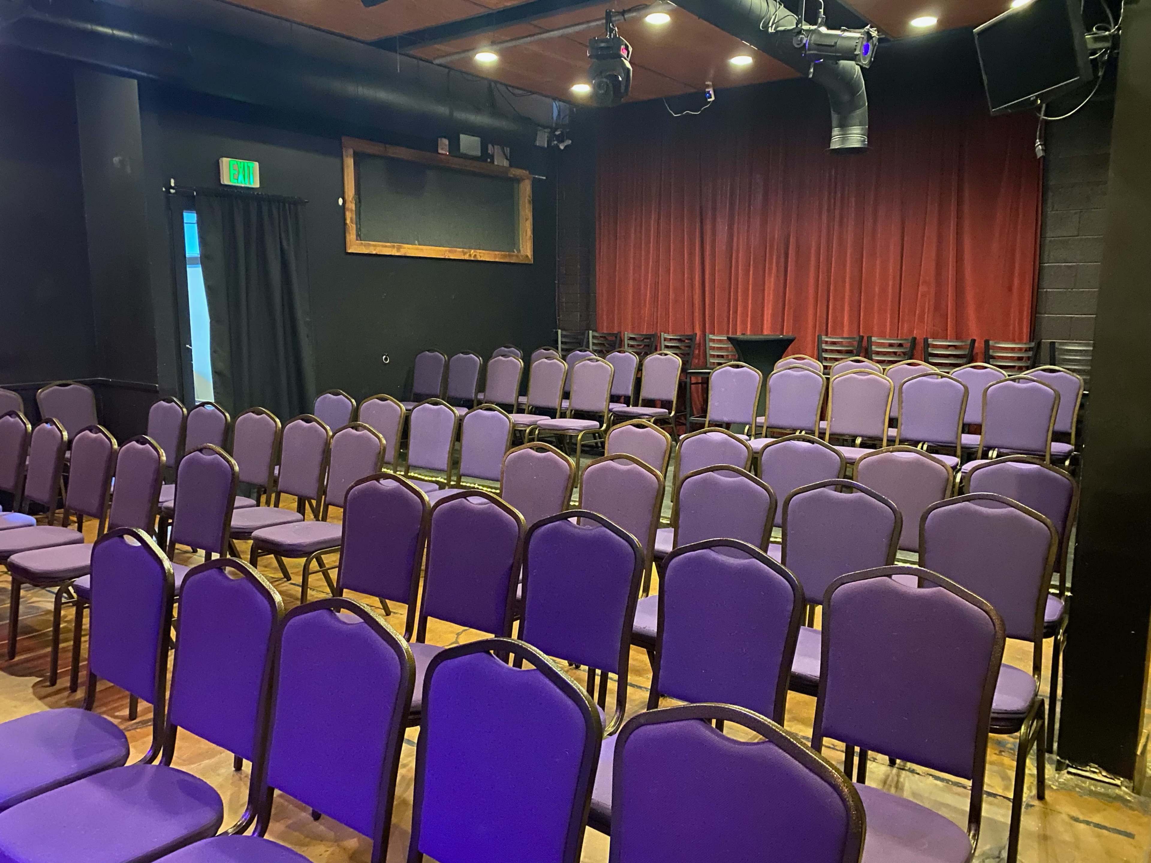 A row of purple chairs is arranged in a small indoor theater space with a stage and curtains in the background.