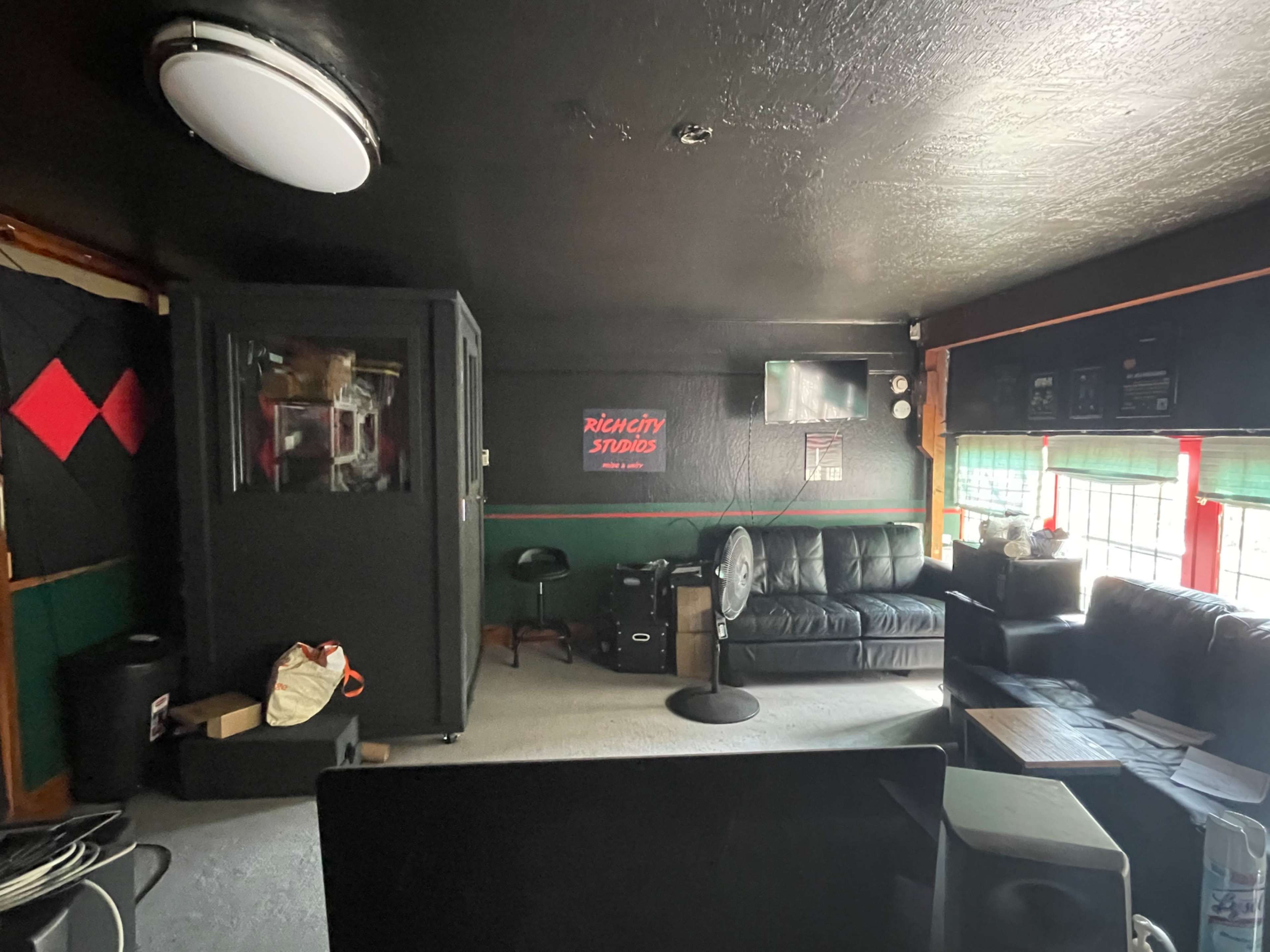 A dimly lit room with a black leather couch, a fan, and a soundproof booth along with a TV on the wall.