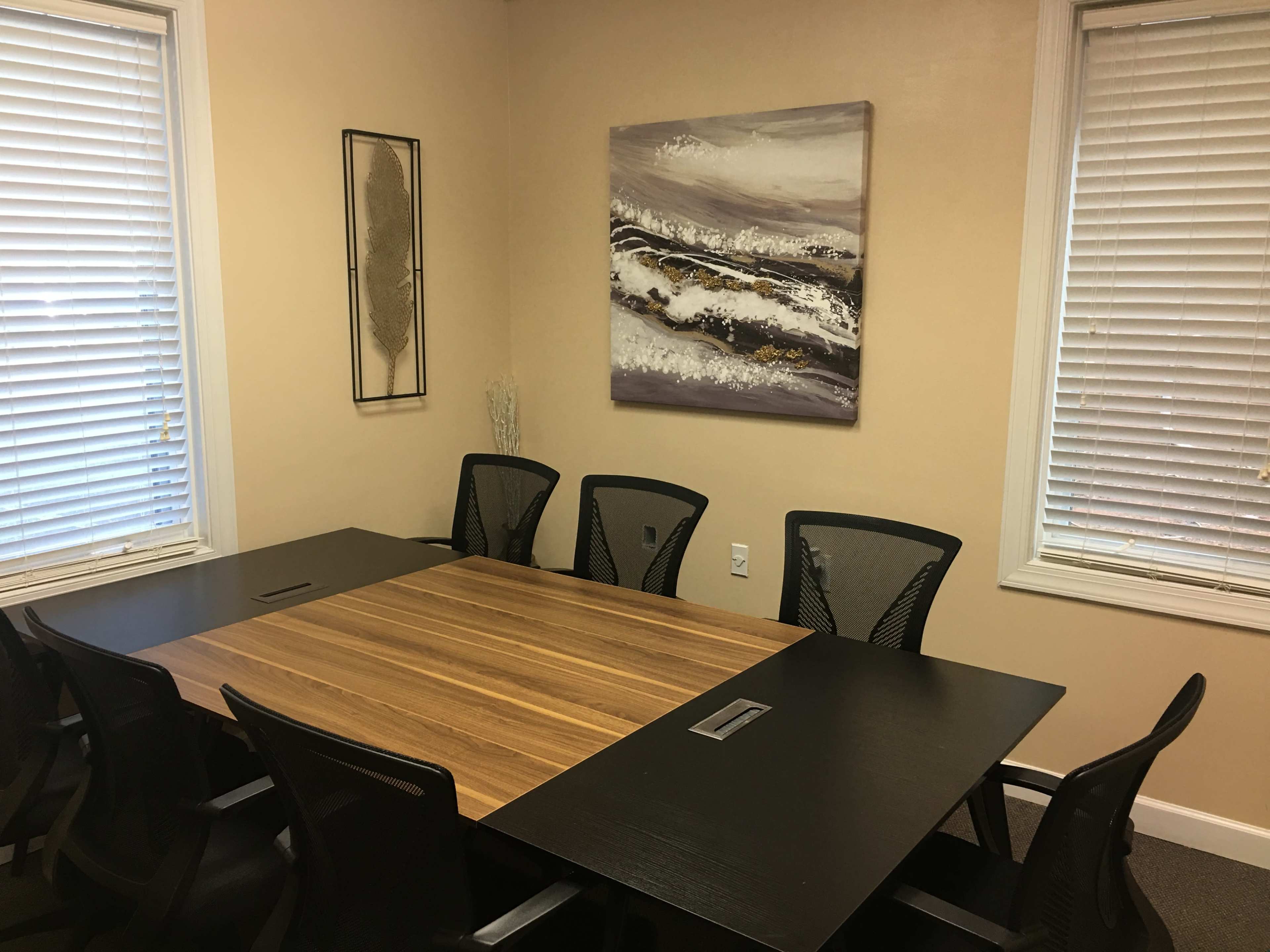 A conference room features a large wooden table surrounded by black mesh chairs, with windows allowing natural light and a landscape painting on the wall.