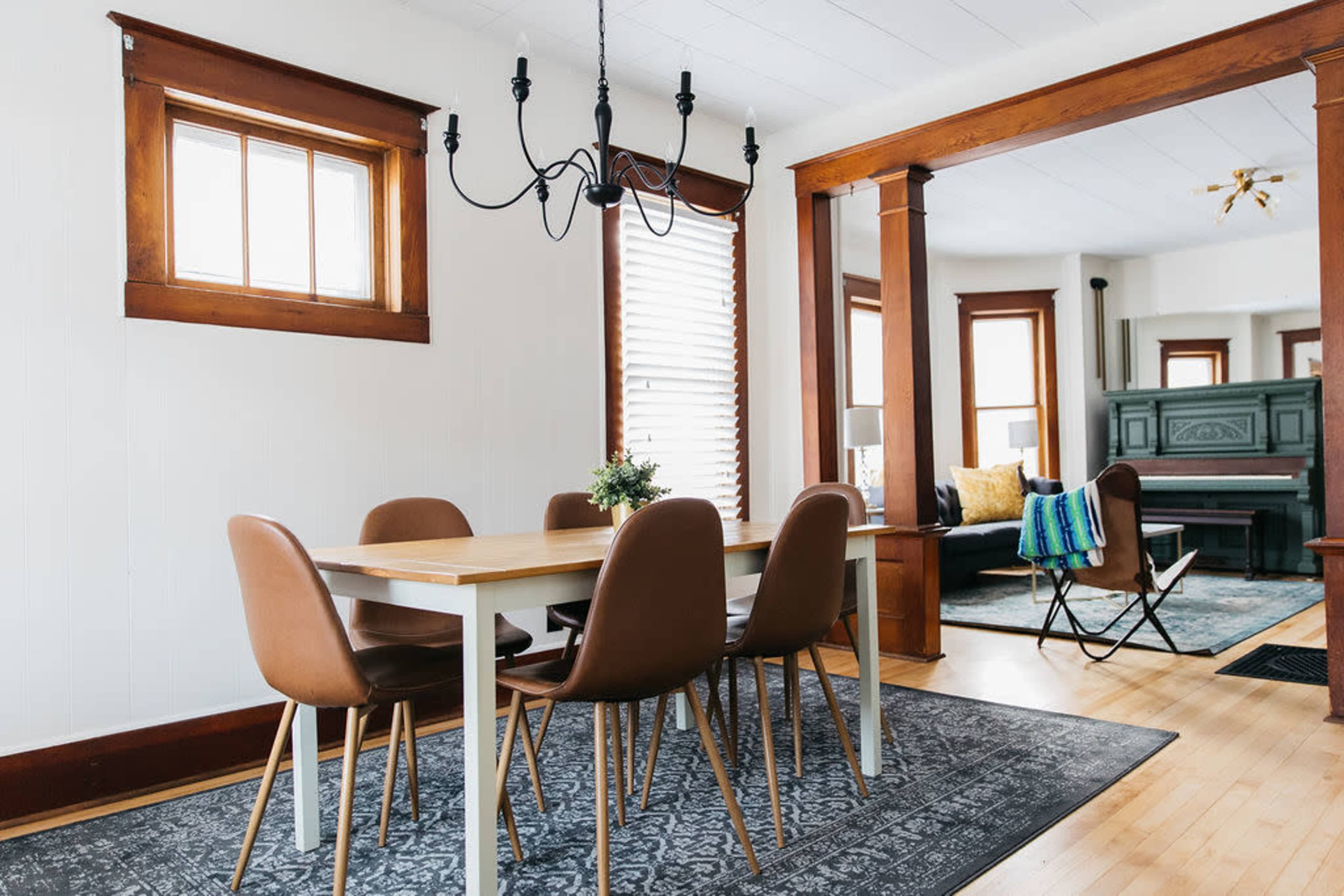 A dining area features a wooden table surrounded by six brown chairs, with a chandelier above and a view into a living room with a piano and seating area.
