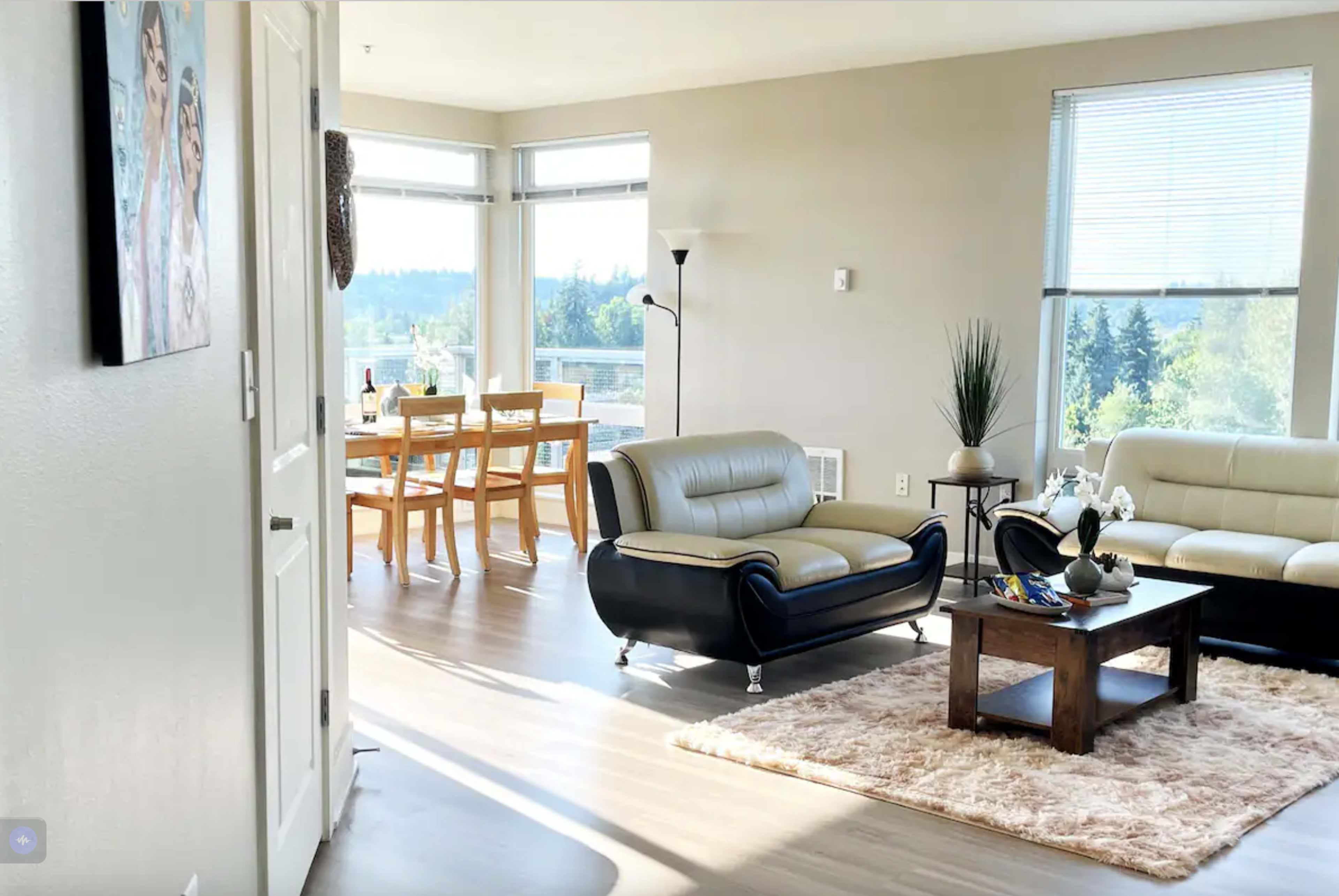 The image depicts a modern living room with two sofas, a coffee table on a plush rug, and a dining area with a table and chairs in bright, natural light.