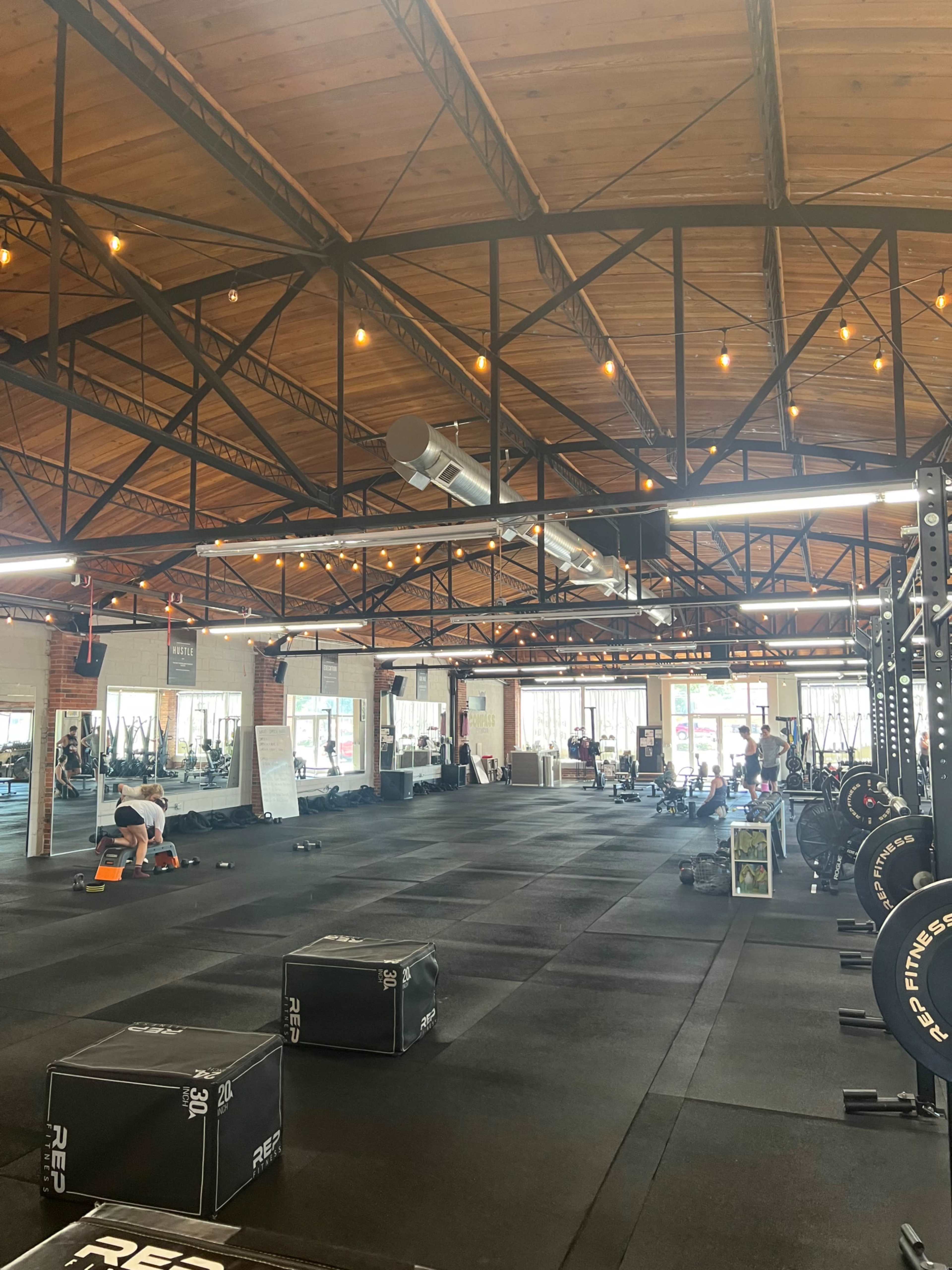 The image shows a spacious gym with exercise equipment, weights, and workout areas under exposed wooden beams and string lights.
