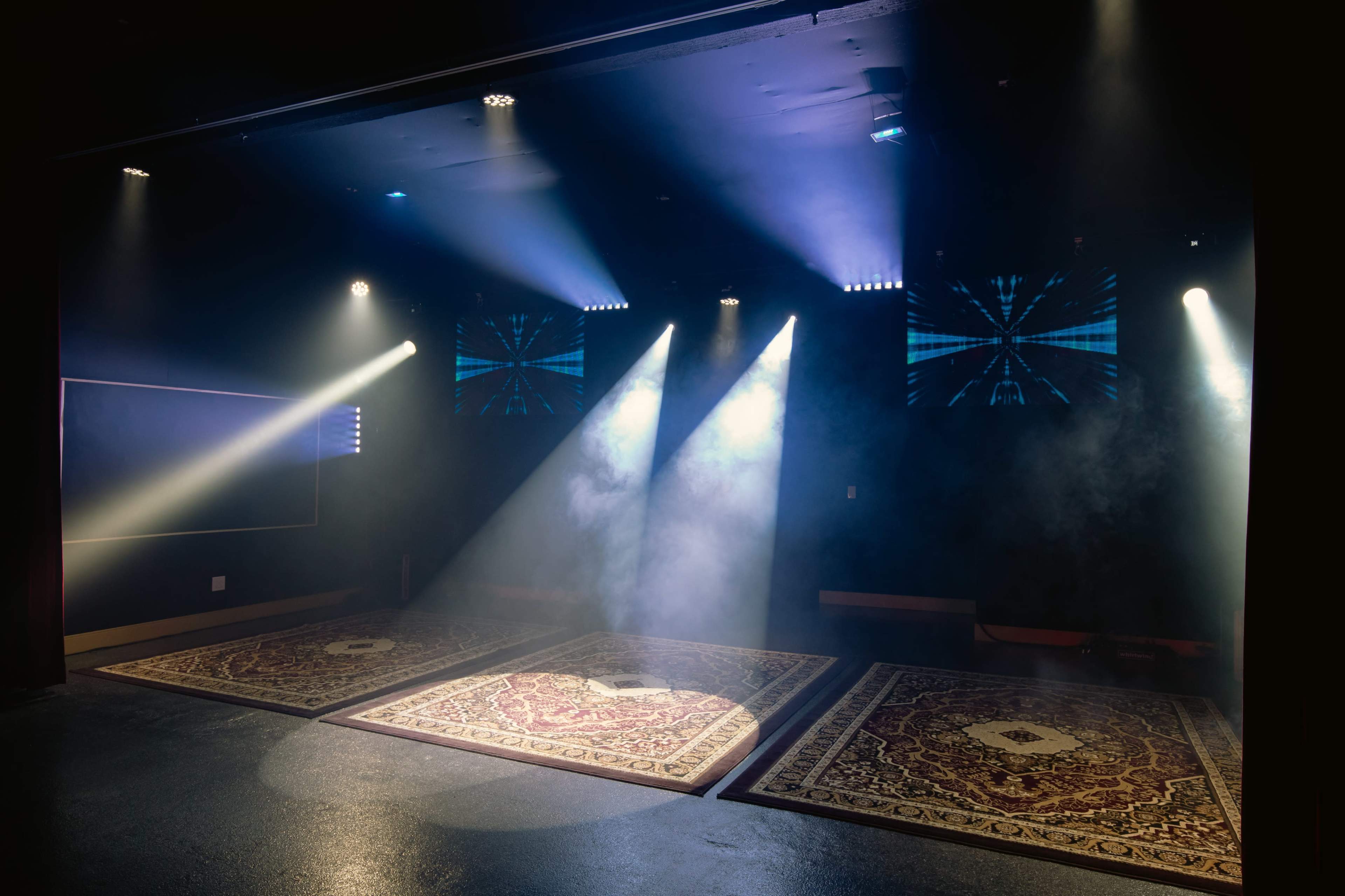 The scene shows a dimly lit stage with two ornate rugs and beams of light cutting through smoke.