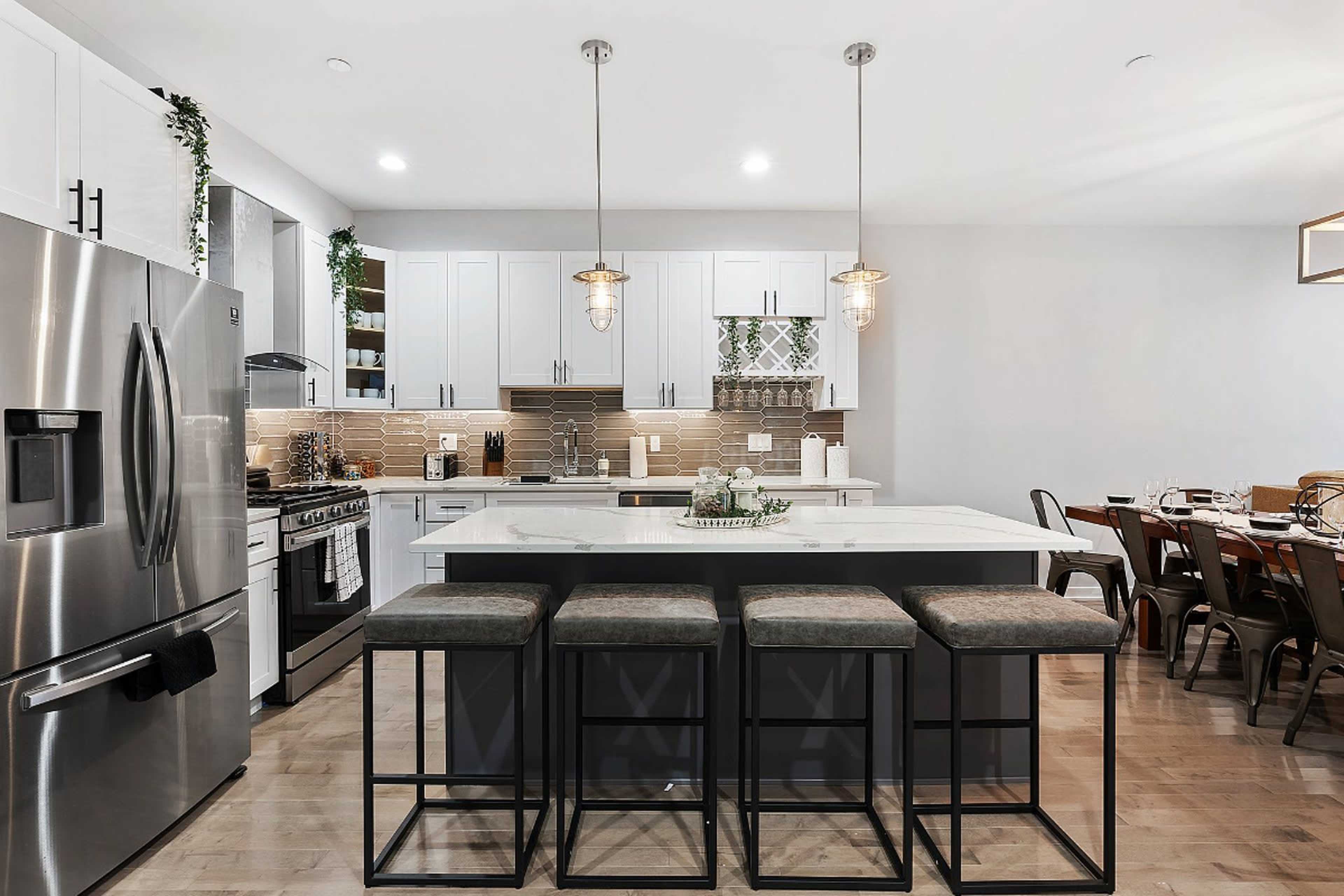 A modern kitchen features white cabinetry, a large island with seating, stainless steel appliances, and pendant lighting.