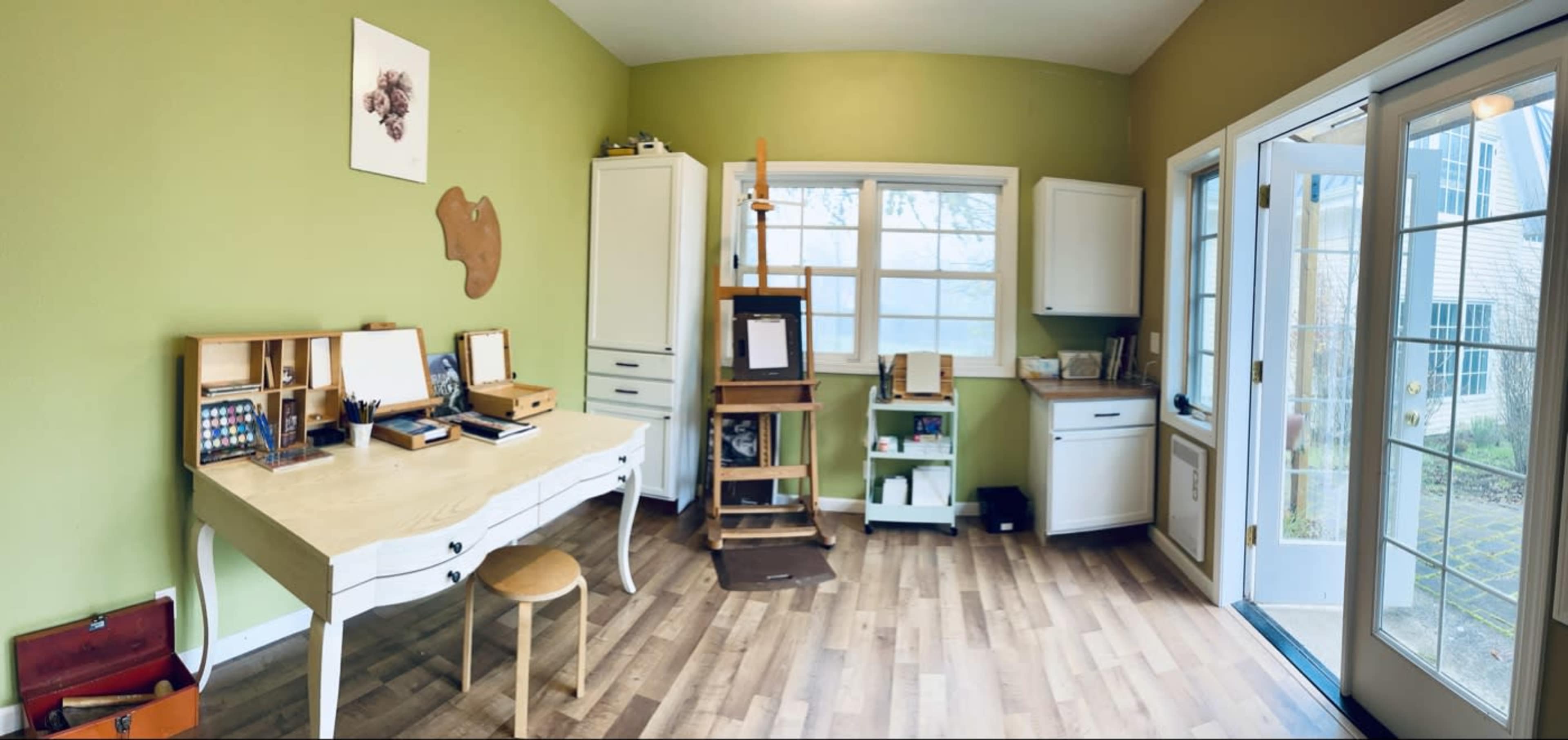 The image depicts a bright green art studio featuring a wooden desk, an easel, various storage cabinets, and large windows letting in natural light.