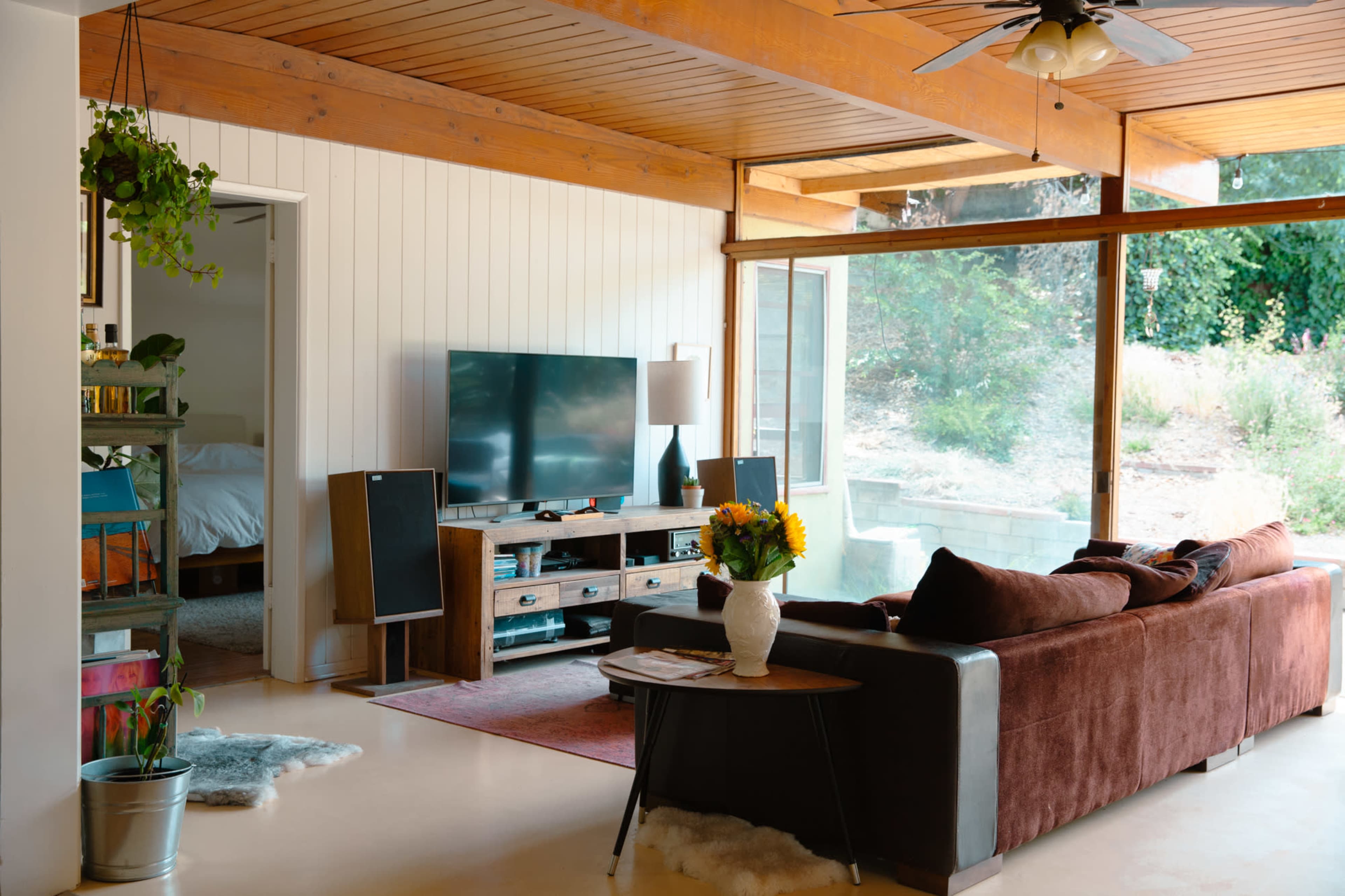 A living room features a brown sofa facing a TV unit, with large windows allowing natural light and a view of the outdoors.
