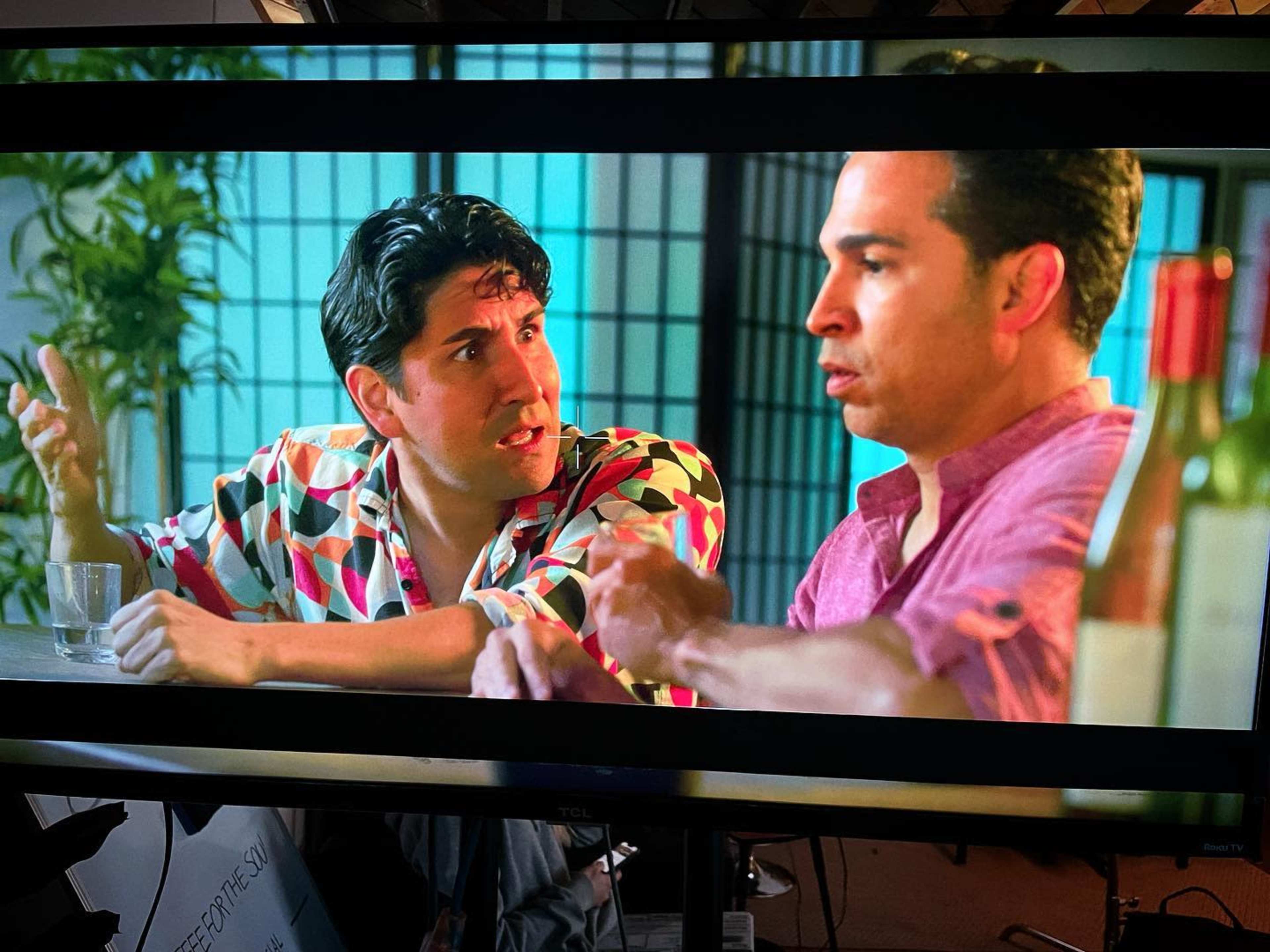 Two men are seated at a table, engaged in an animated conversation, with bottles and glasses visible nearby.