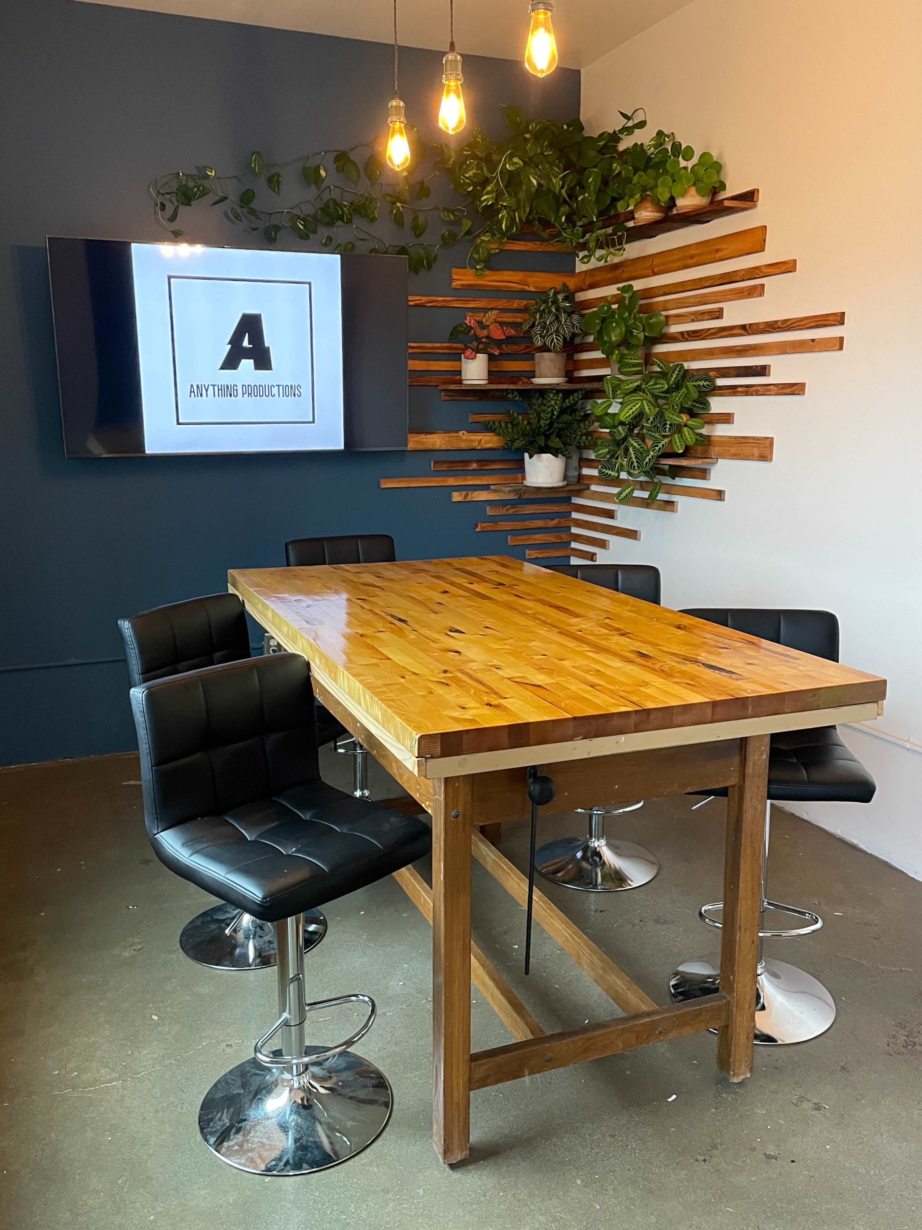 A modern meeting room features a wooden table surrounded by black leather chairs, with a wall adorned by plants and a television displaying the text "Anything Productive."