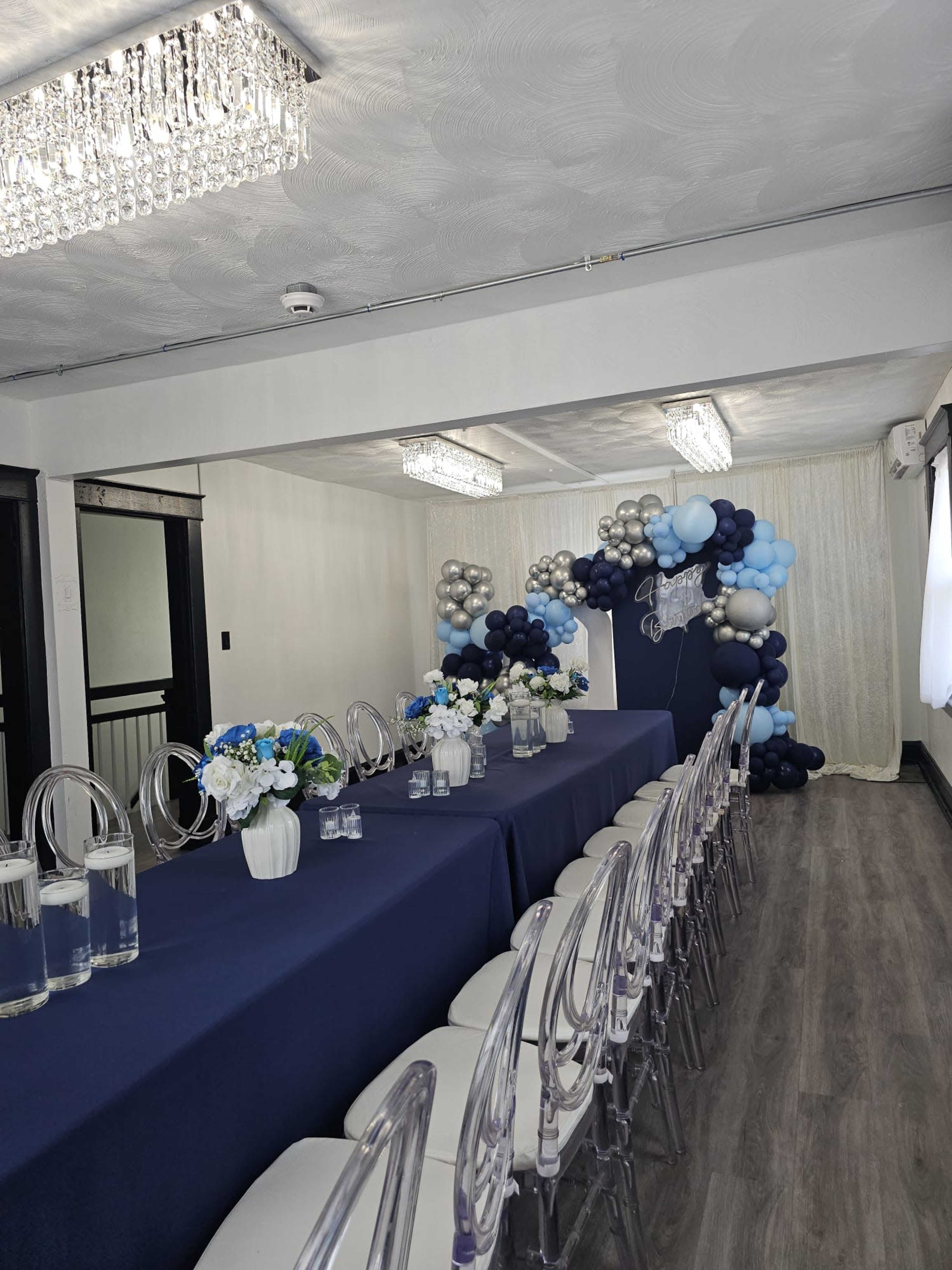 The image shows a decorated indoor event space featuring a long table set with clear chairs, floral centerpieces, and a backdrop of blue, gray, and silver balloons.