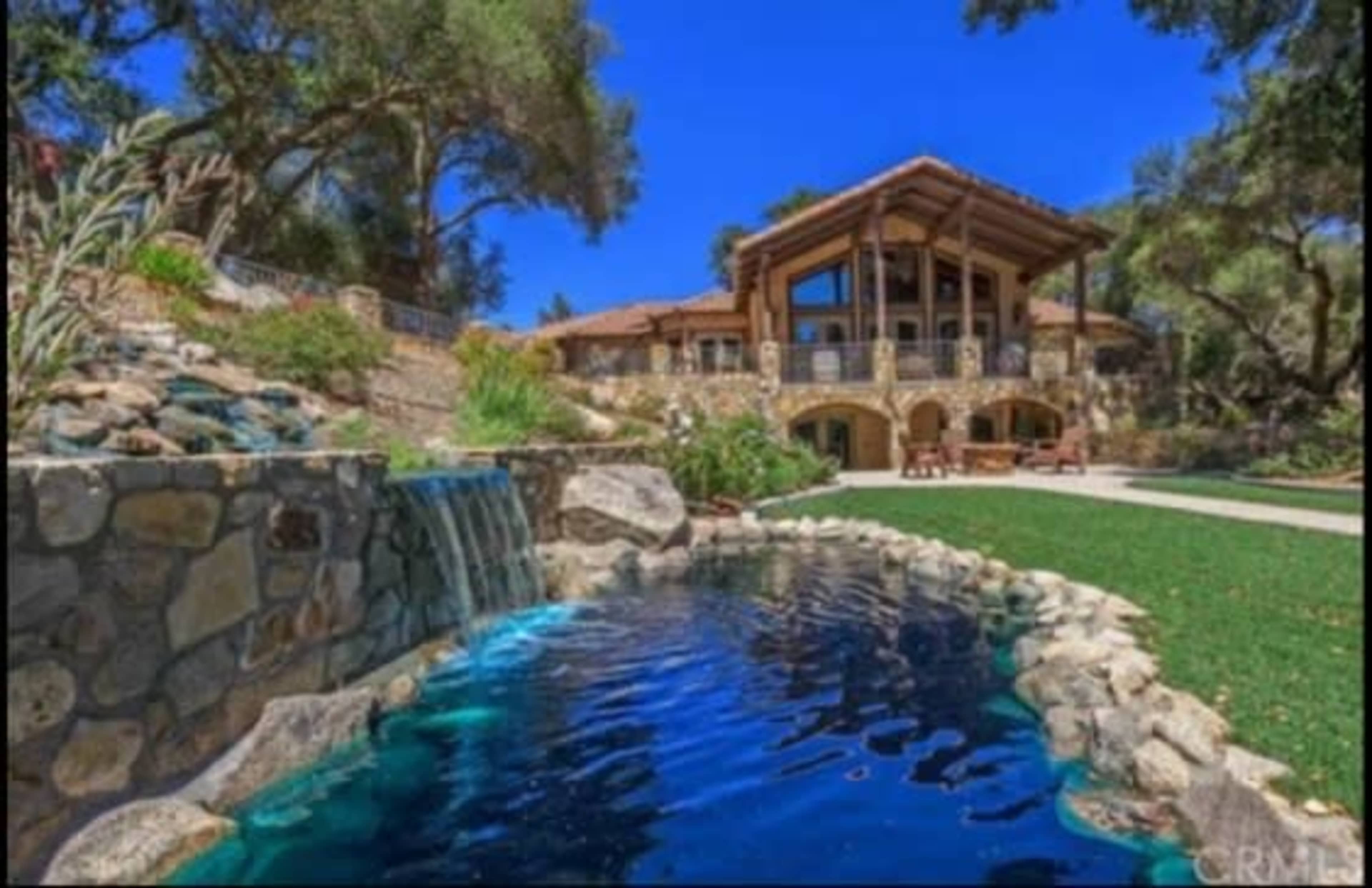 A large house with wooden elements, surrounded by a landscaped yard featuring a stone waterfall and a clear blue pond.